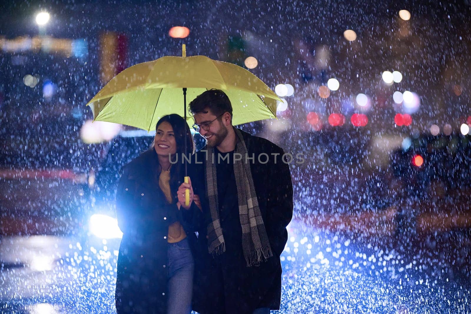 In the romantic ambiance of a rainy night, a happy couple walks through the city, sharing tender moments under a yellow umbrella, surrounded by the glistening glow of urban lights.