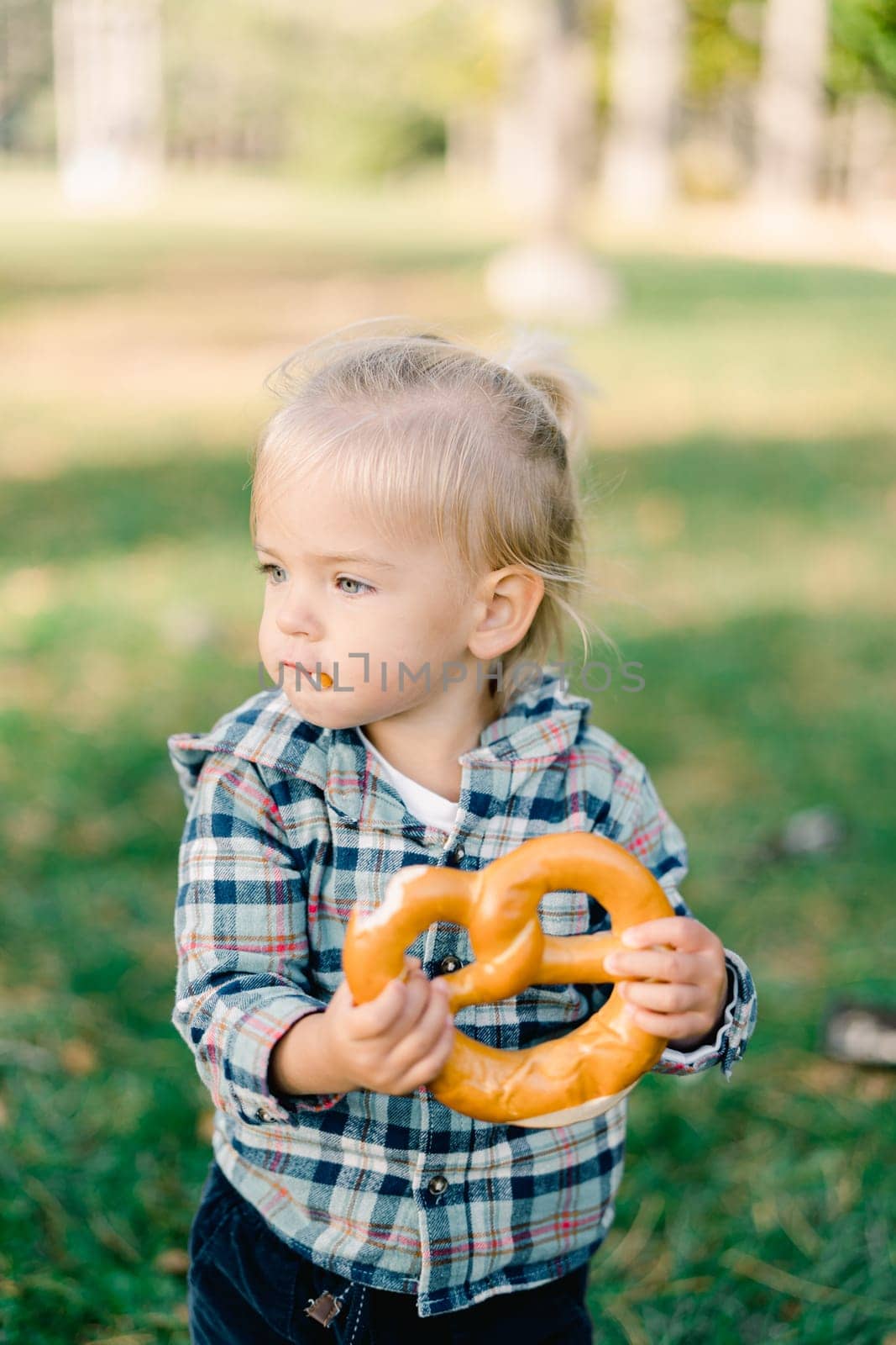 Little girl is eating a big bagel, holding it with both hands and turning her head to the side. High quality photo