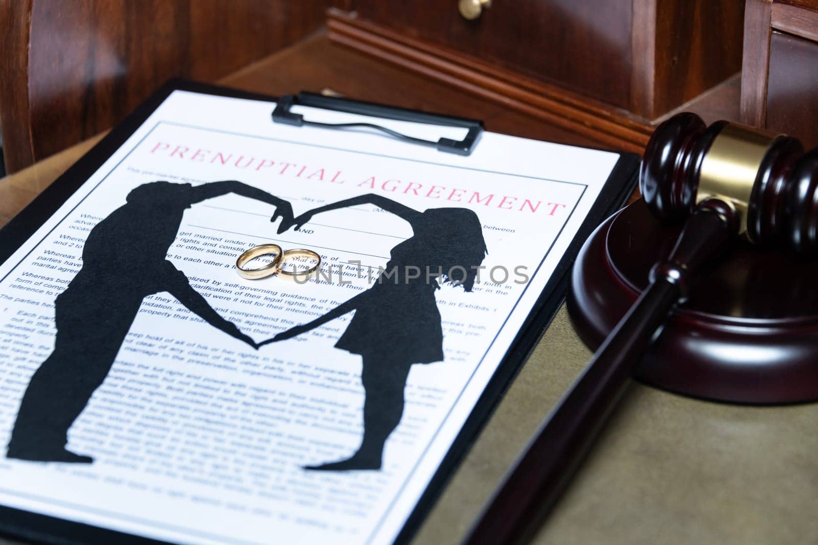 A clipboard holding a prenuptial agreement with a cut-out silhouette of a couple and wedding rings, next to a judge's gavel