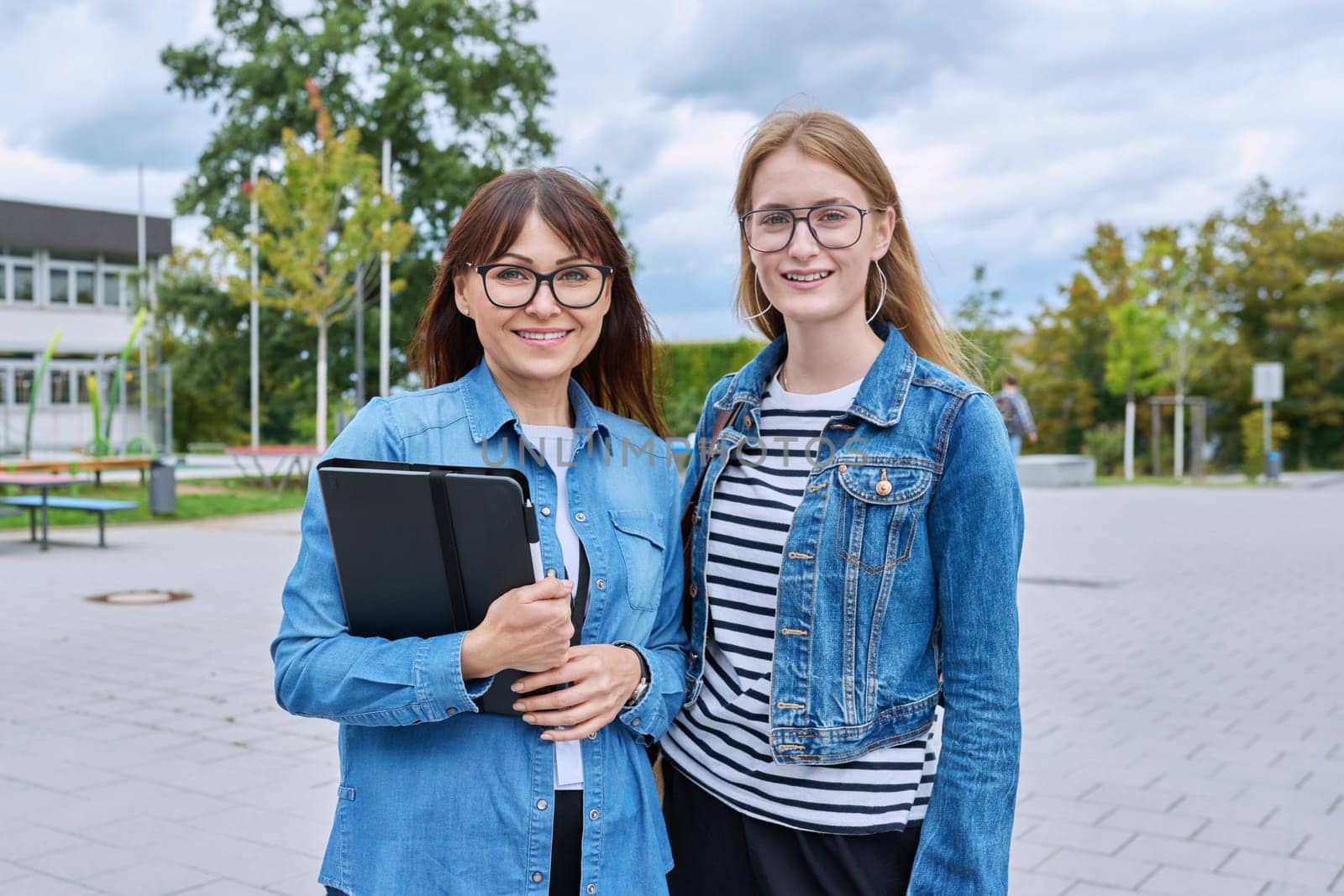 Teacher and teenage schoolgirl looking at camera together outdoor, school building background. Meeting communication student girl with backpack and mentor counselor. Education, adolescence, learning concept