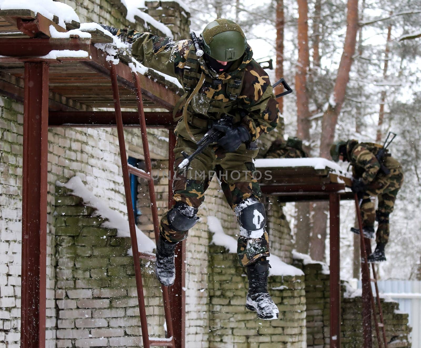 Special forces soldiers climbing a building in combat gear during a military exercise. Armed with rifles, helmets, and protective gear. Brick building with windows in the woods.