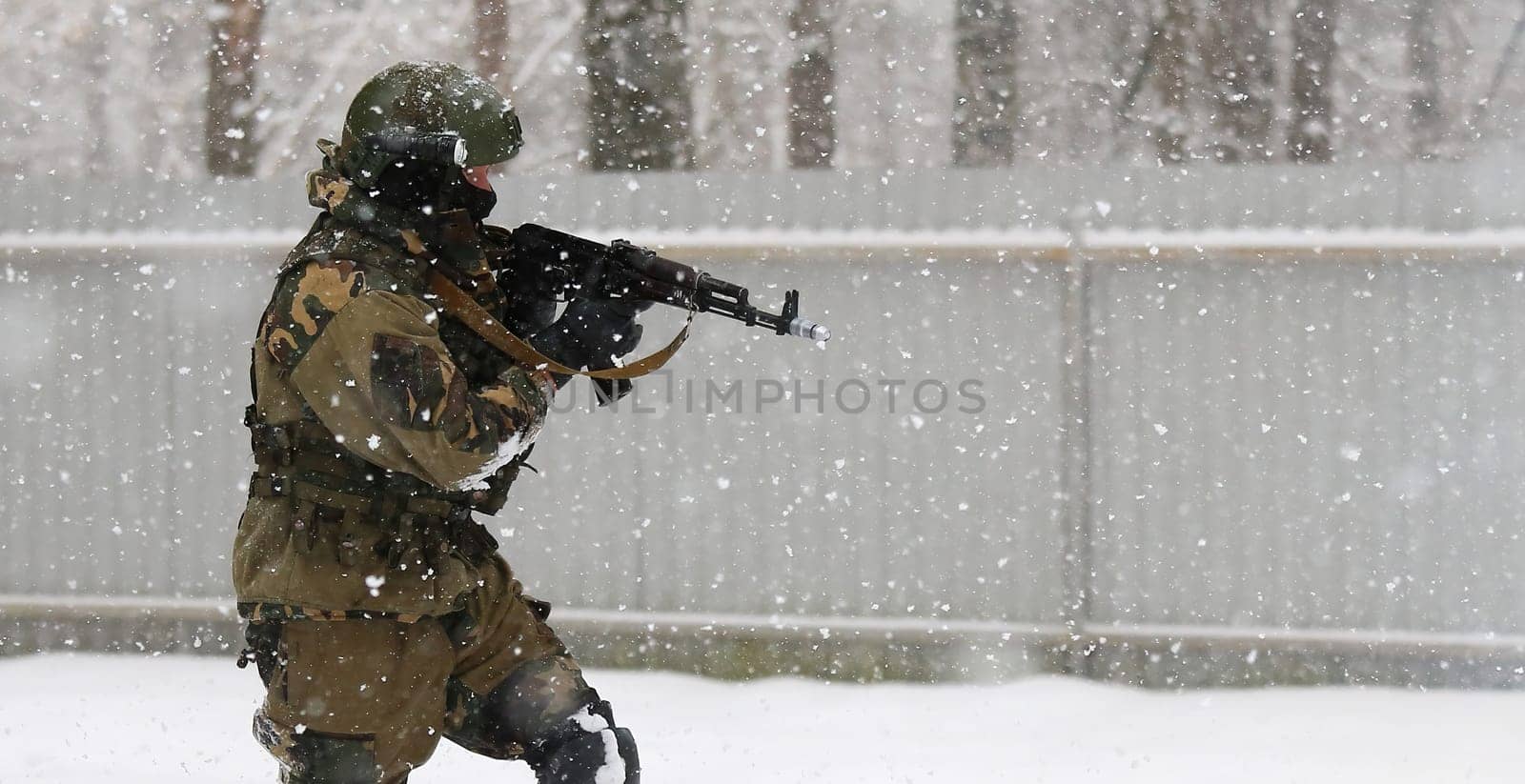 A soldier wearing a mask and winter camouflage uniform is pictured advancing through the snow while holding a gun. The soldier is likely participating in a military operation or training exercise.