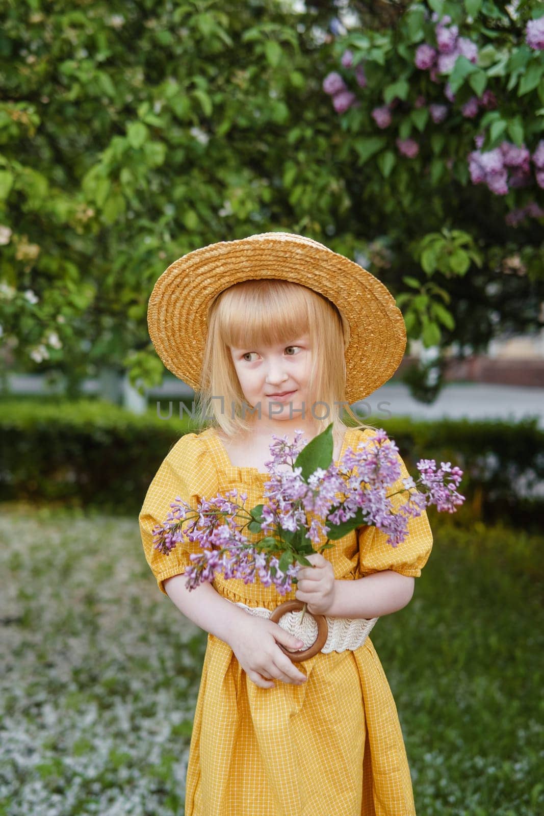 A little girl in a yellow dress and straw hat wearing a bouquet of lilacs. A walk in a spring park, blossoming lilacs