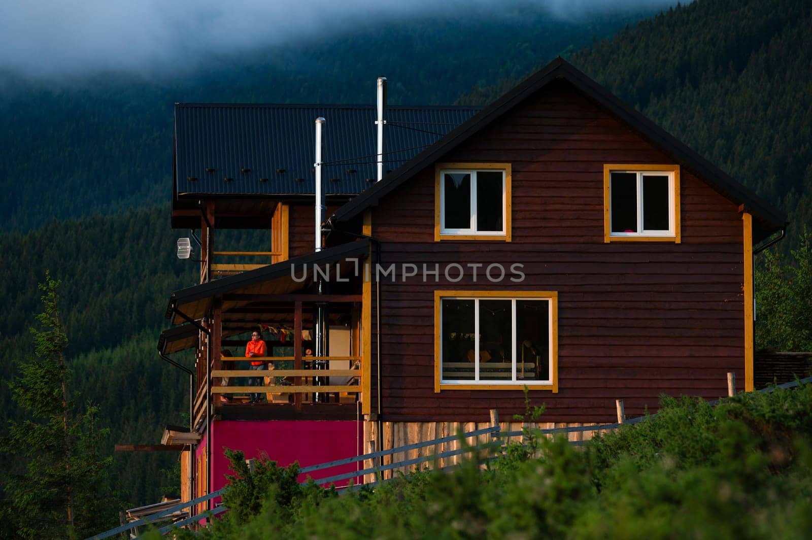 a girl in an orange jacket stands on the terrace of a wooden house against the background of mountains by Niko_Cingaryuk