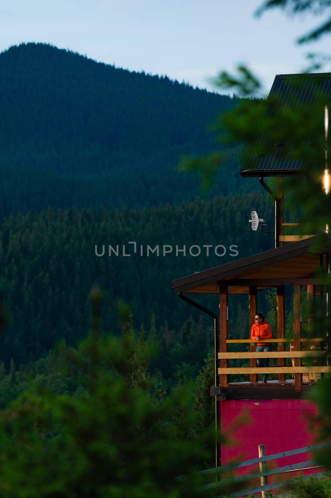 a girl in an orange jacket stands on the terrace of a wooden house against the background of mountains, a house for tourists in the Carpathians of Ukraine.