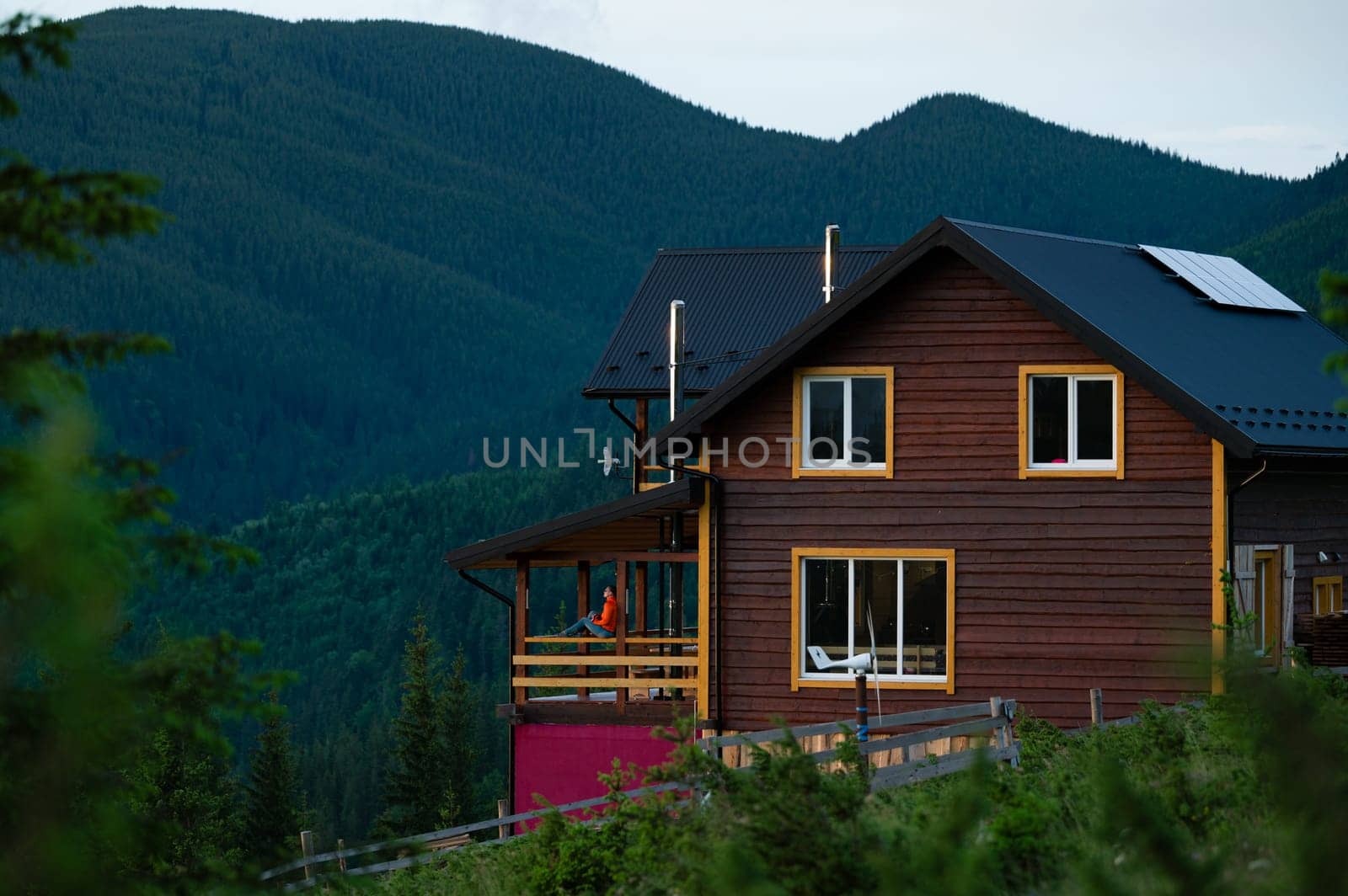A large wooden house for tourists in the mountains, a young girl stands on the terrace against the background of the Carpathian mountains. by Niko_Cingaryuk