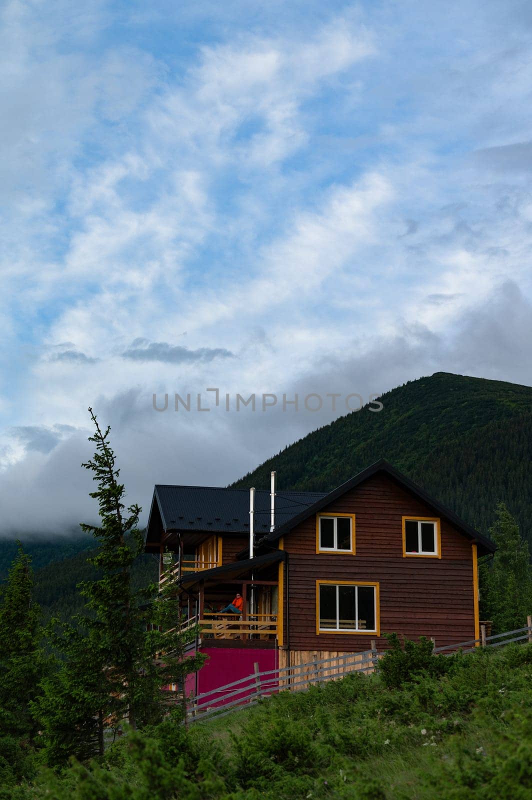 A large wooden house for tourists in the mountains, a young girl stands on the terrace against the background of the Carpathian Mountains, a house for recreation in the mountains.