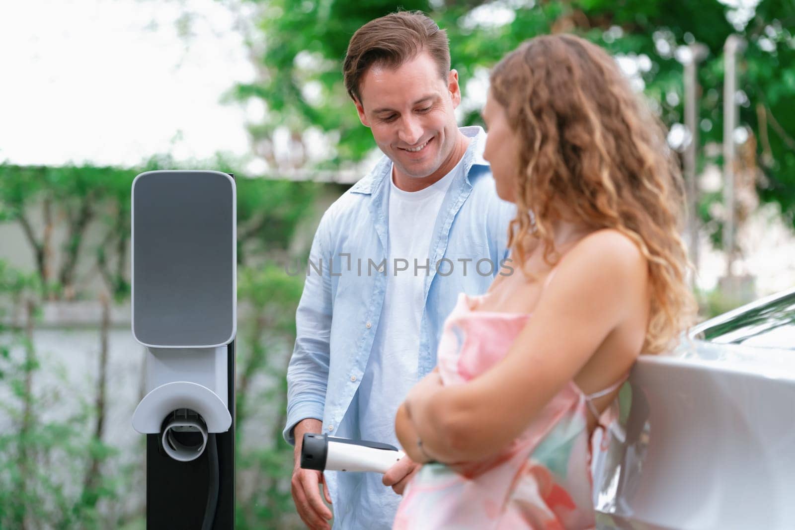 Happy and lovely couple with eco-friendly conscious recharging electric vehicle from EV charging station. EV car technology utilized as alternative transportation for future sustainability. Synchronos