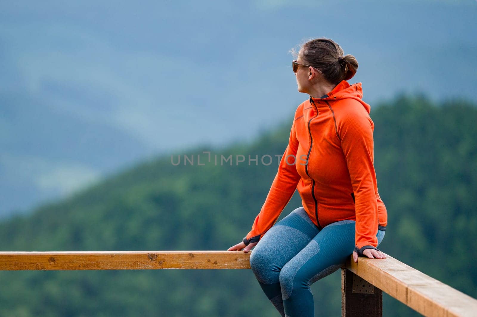 a young girl sits on a wooden handrail, against the background of mountains and evergreen forest.