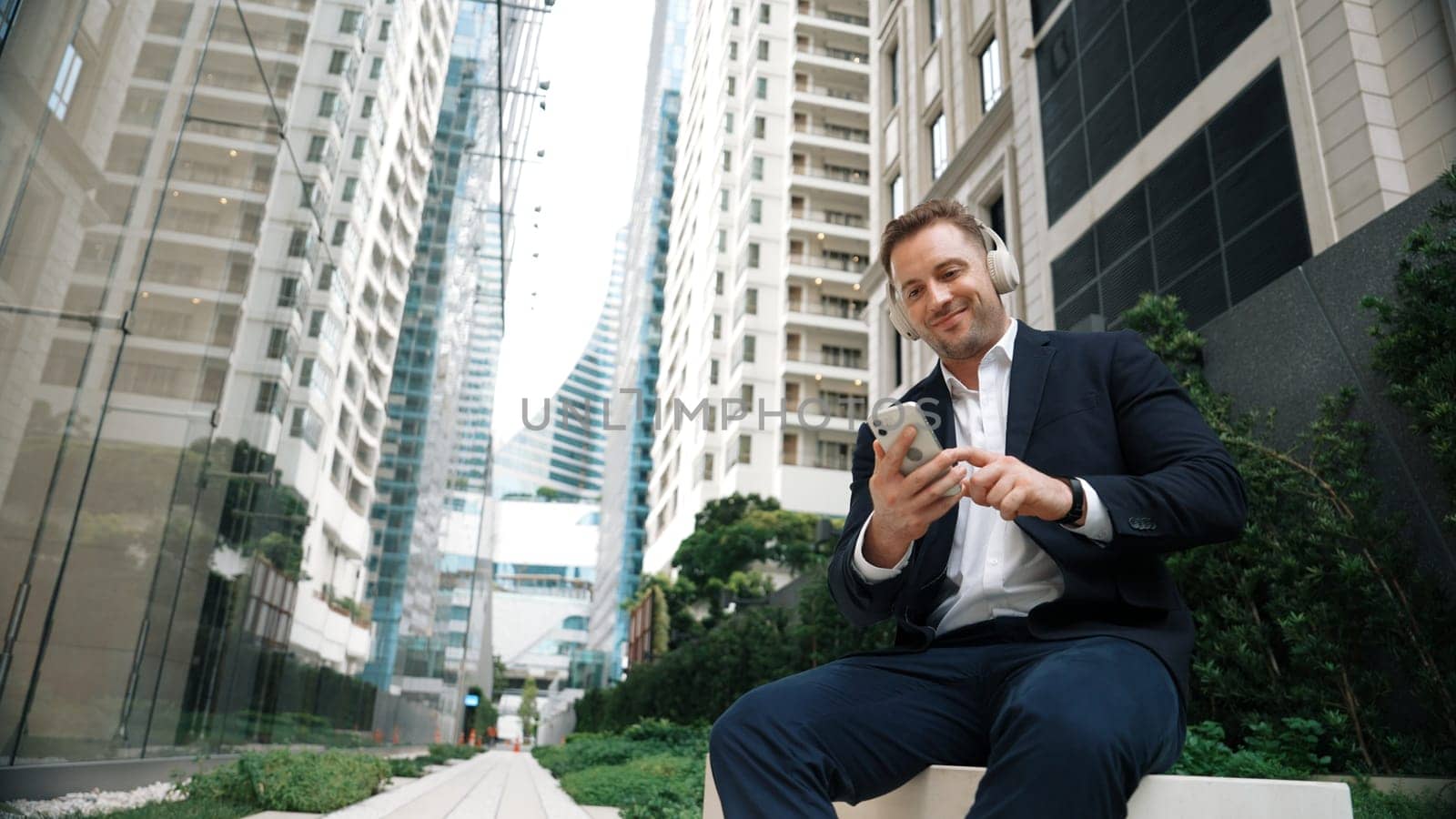 Caucasian businessman listening relaxing music while hold mobile phone. Skilled happy executive manager open music and move to song in lively mood while sitting at green urban city or park. Urbane.