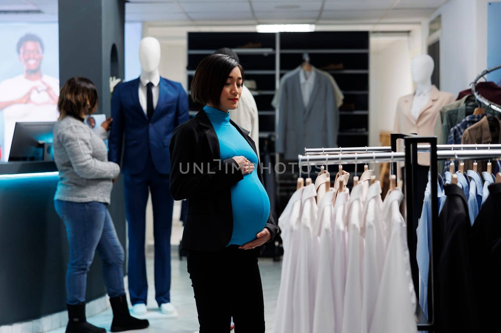 Pregnant asian woman exploring clothing store and searching for maternity apparel. Expectant mother holding belly while standing near garment rack in retail market boutique