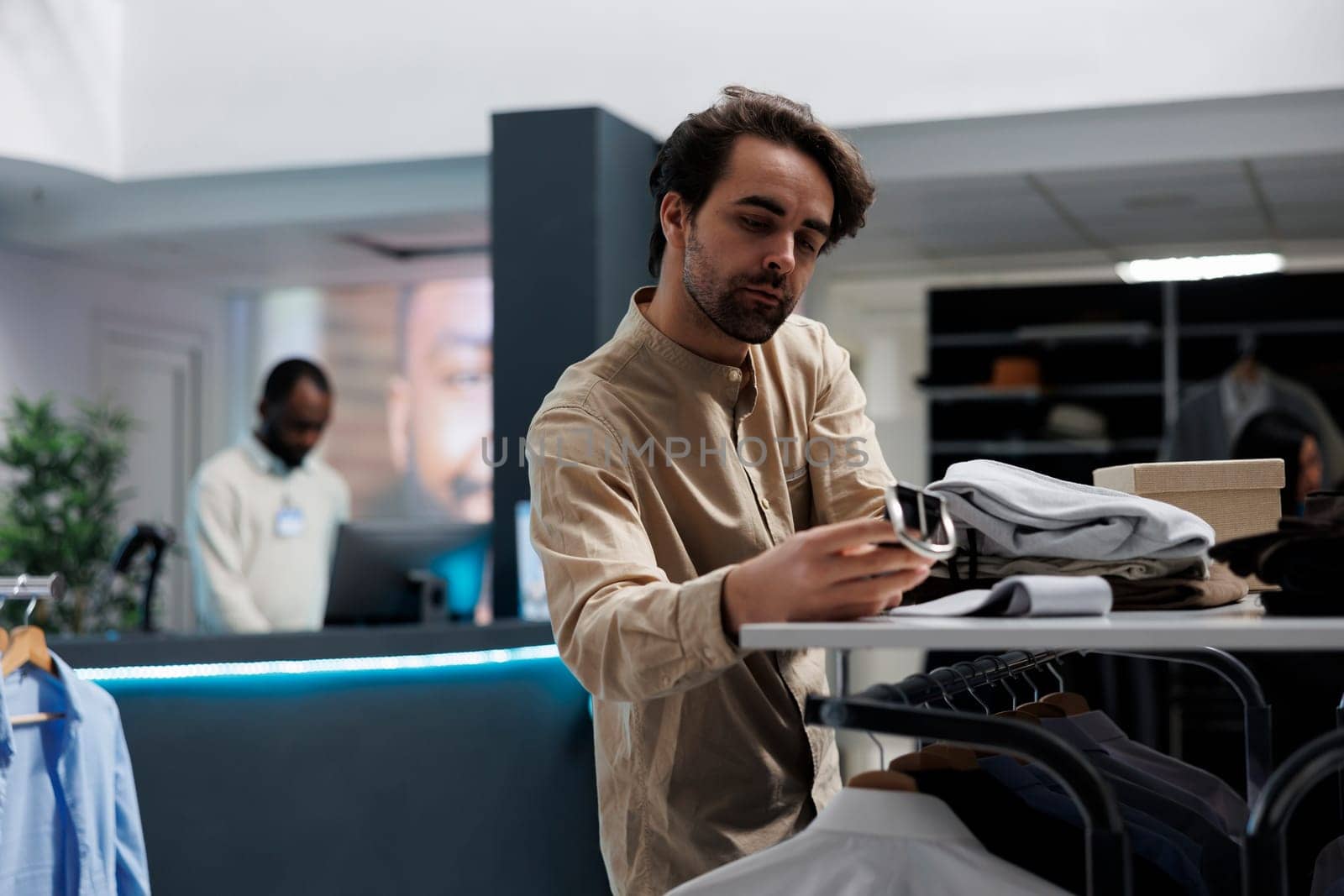 Clothing store client examining belt style and quality before making purchase. Young man searching for formal outfit, browsing shelf and selecting accessory in menswear showroom