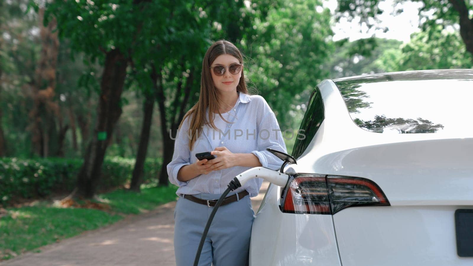 Woman using smartphone online banking application to pay for electric car battery charging from EV charging station during vacation holiday road trip at national park or summer forest. Exalt