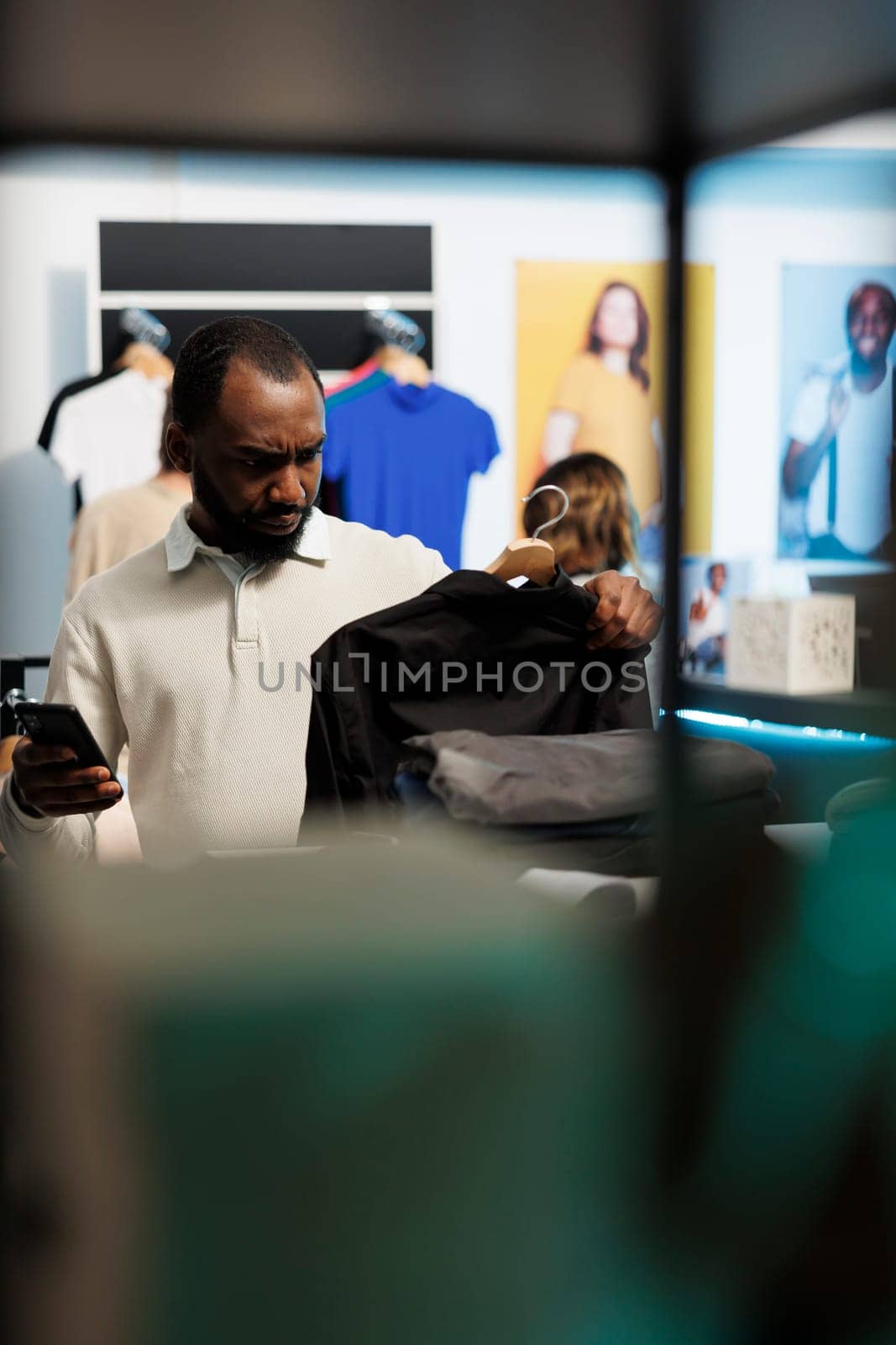 African american man shopping for shirt and checking apparel in stock using smartphone online app. Clothing store customer holding outfit on rack and scrolling website on mobile phone