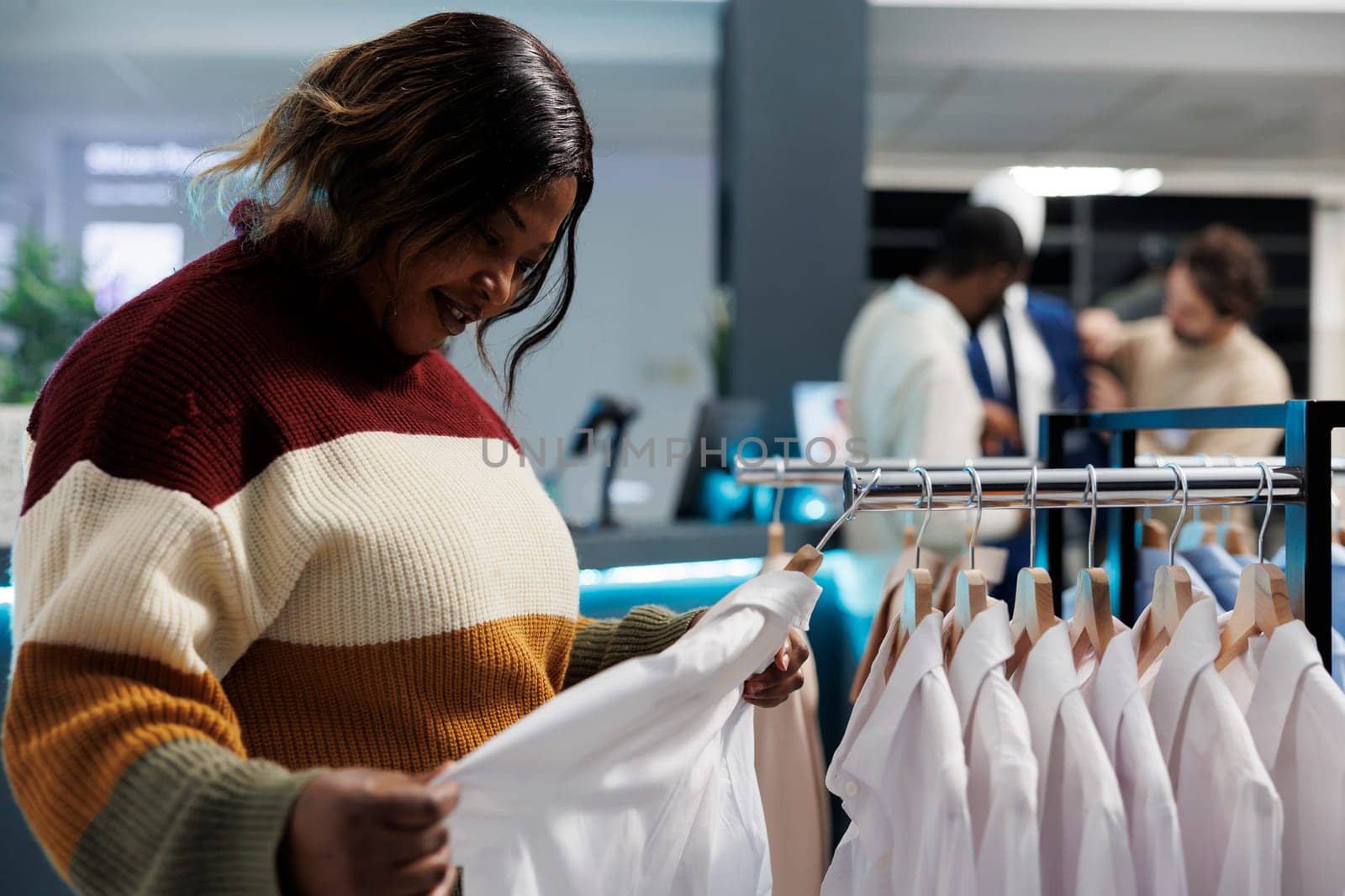 Clothing store buyer examining white shirt fabric quality, checking fit. African american woman browsing rack with hanging formal wear and selecting apparel size in boutique