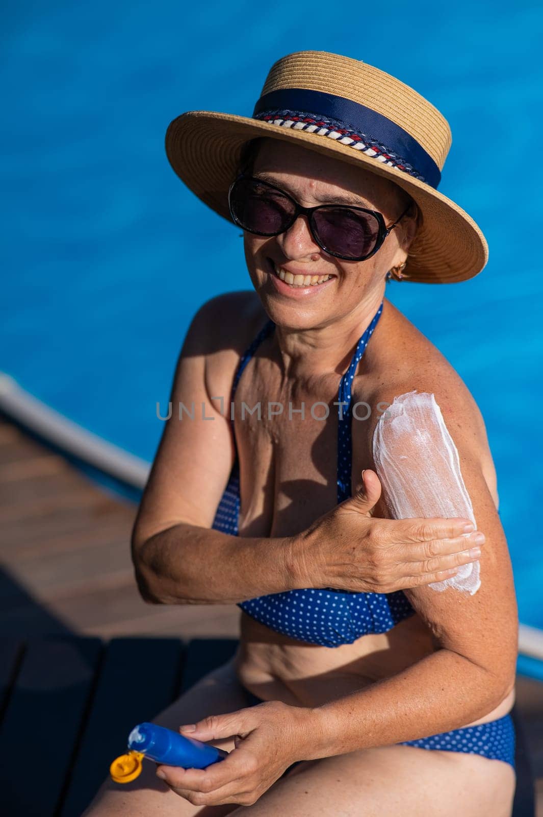 Portrait of an old woman in a straw hat, sunglasses and a swimsuit applying sunscreen to her skin while relaxing by the pool