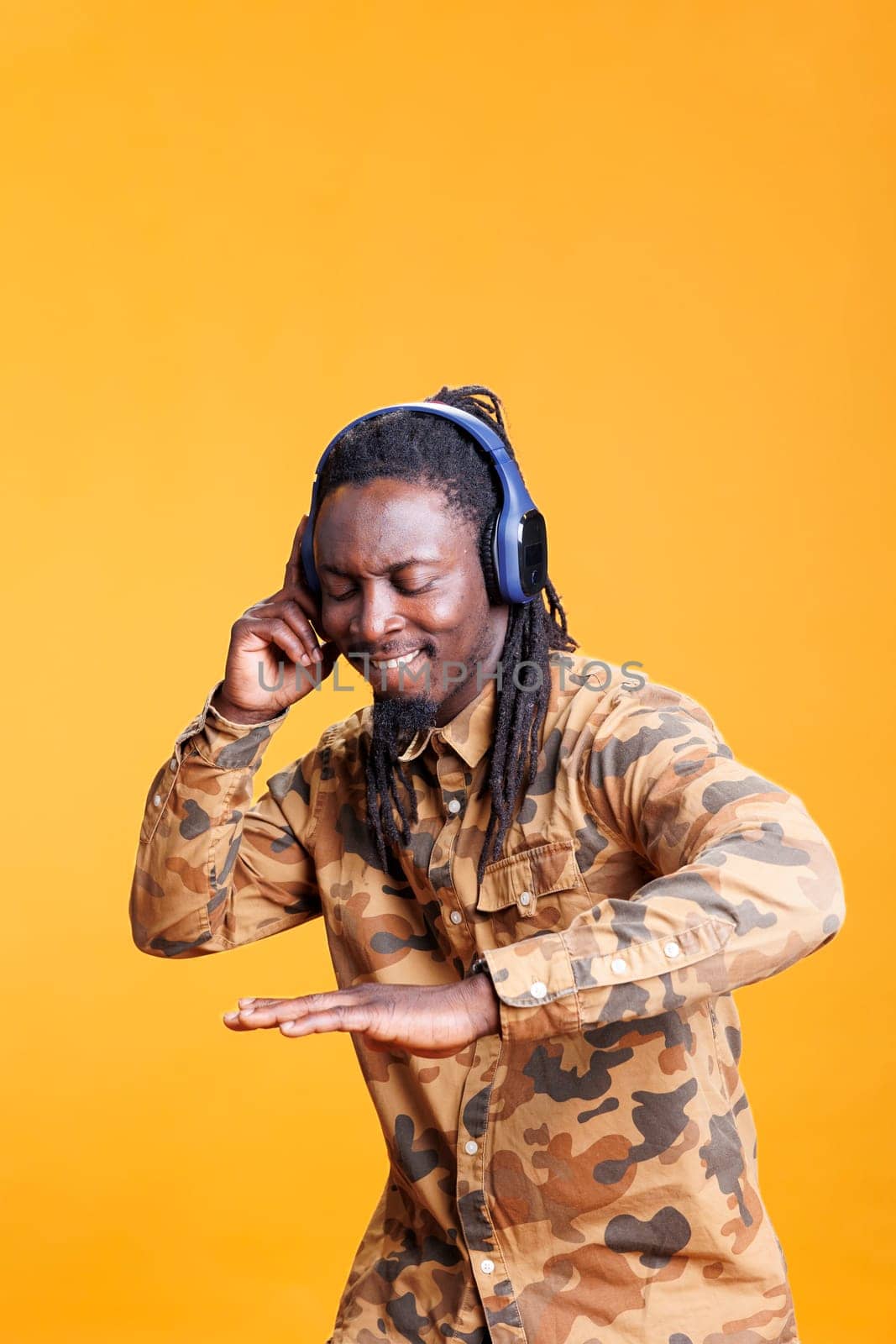 Smiling cheerful man enjoying song on headphones in studio over yellow background, dancing and having fun during leisure time. African american person relaxing and listening to audio on camera