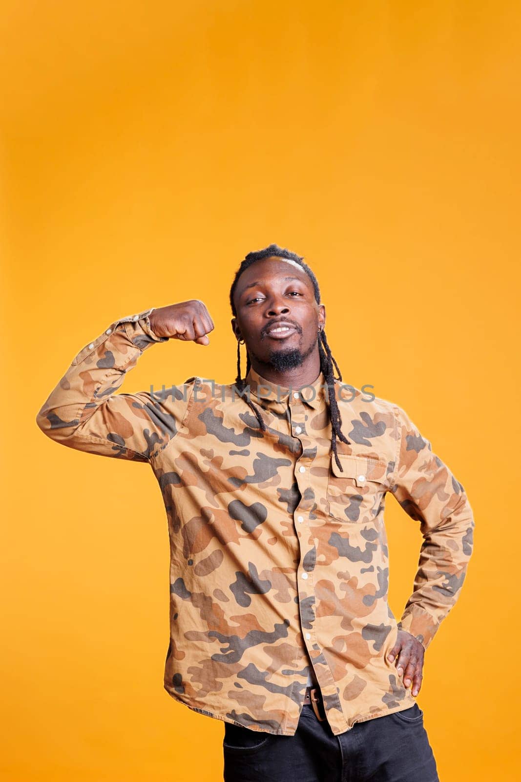 Portrait of athletic bodybuilder showing trained arm muscles in front of camera in studio over yellow background. African american man enjoying sport exercices, working out at triceps strength.