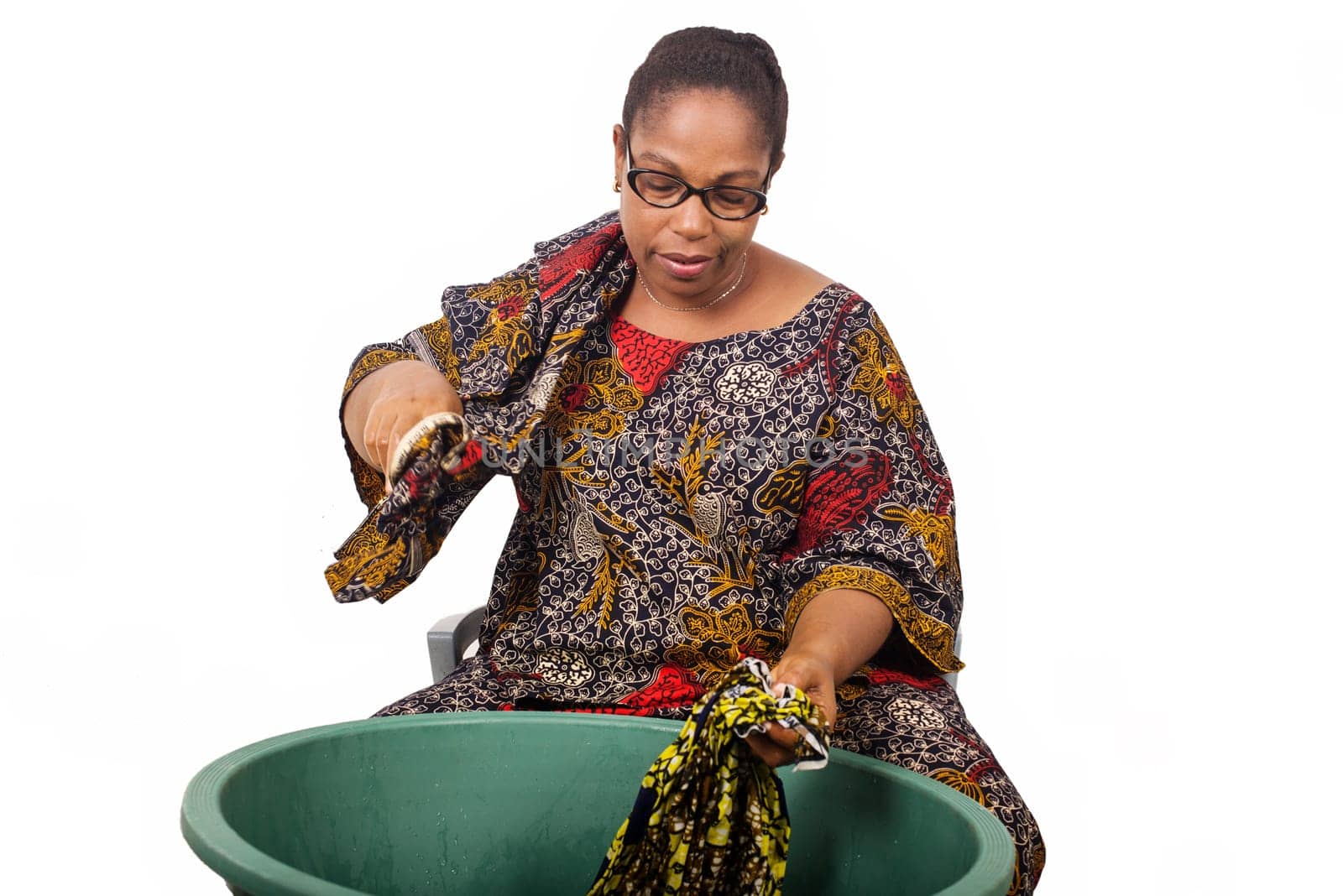 African woman sitting and washing clothes in a big green bowl while arranging her scarf on the shoulder, isolated on white background.