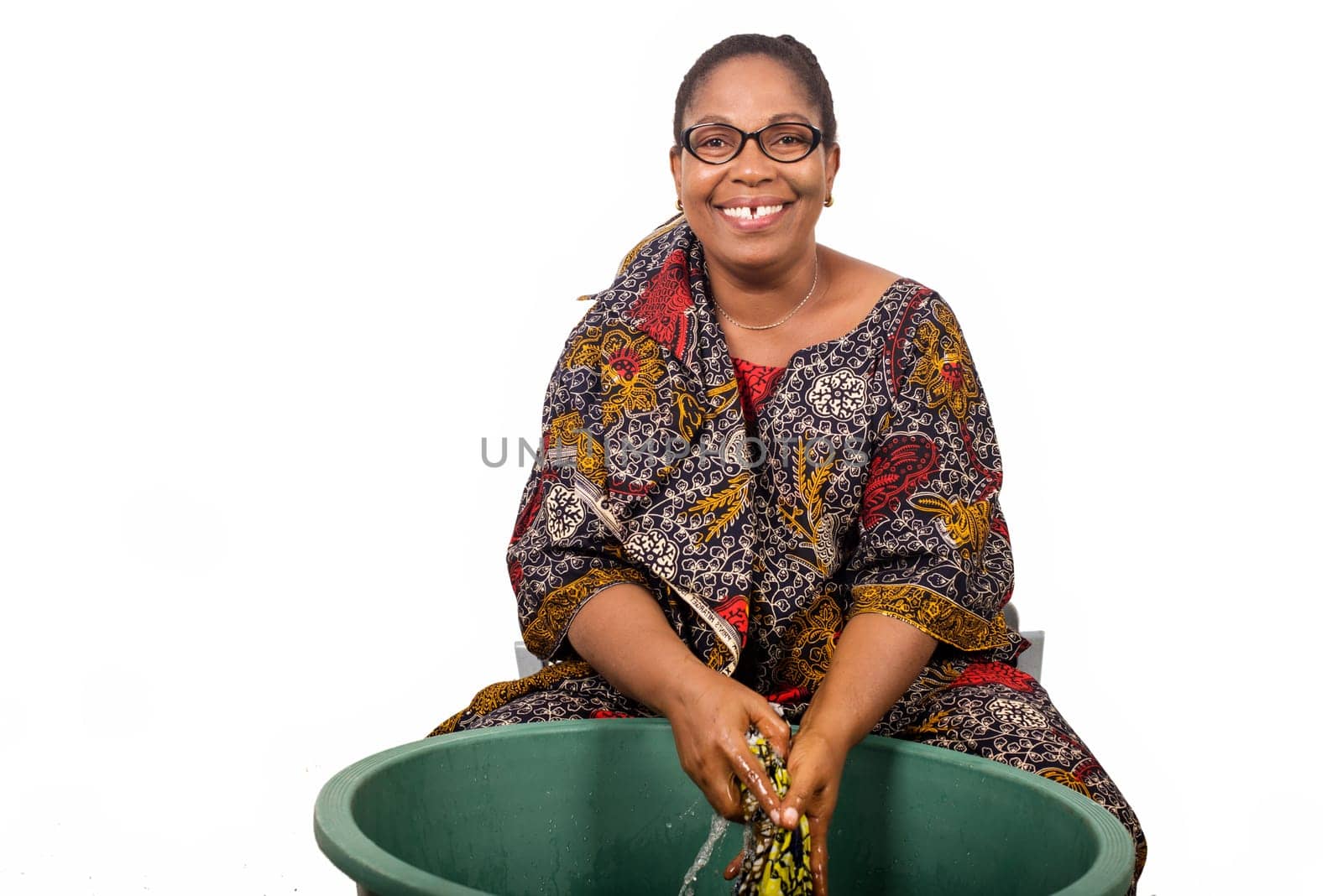 smiling african woman sitting and washing clothes in a big green bowl isolated on white background.