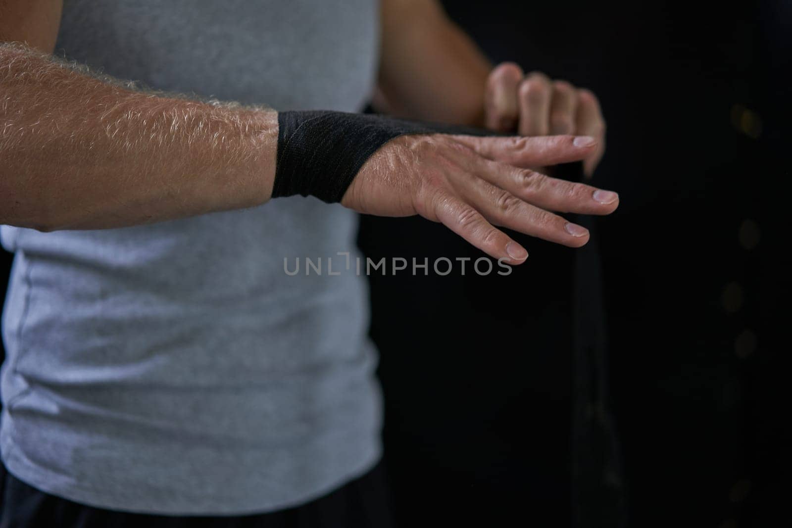 Crop unrecognizable male kickboxer wrapping bandage before training. Sportsman wearing black tape on hand against dark background.