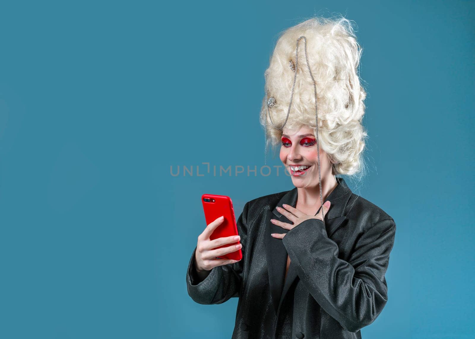 Cheerful young woman in red makeup and royal vintage wig using smartphone. Amazed funny princess wearing black jacket looking at screen of cellphone. Happy lady standing on blue background.