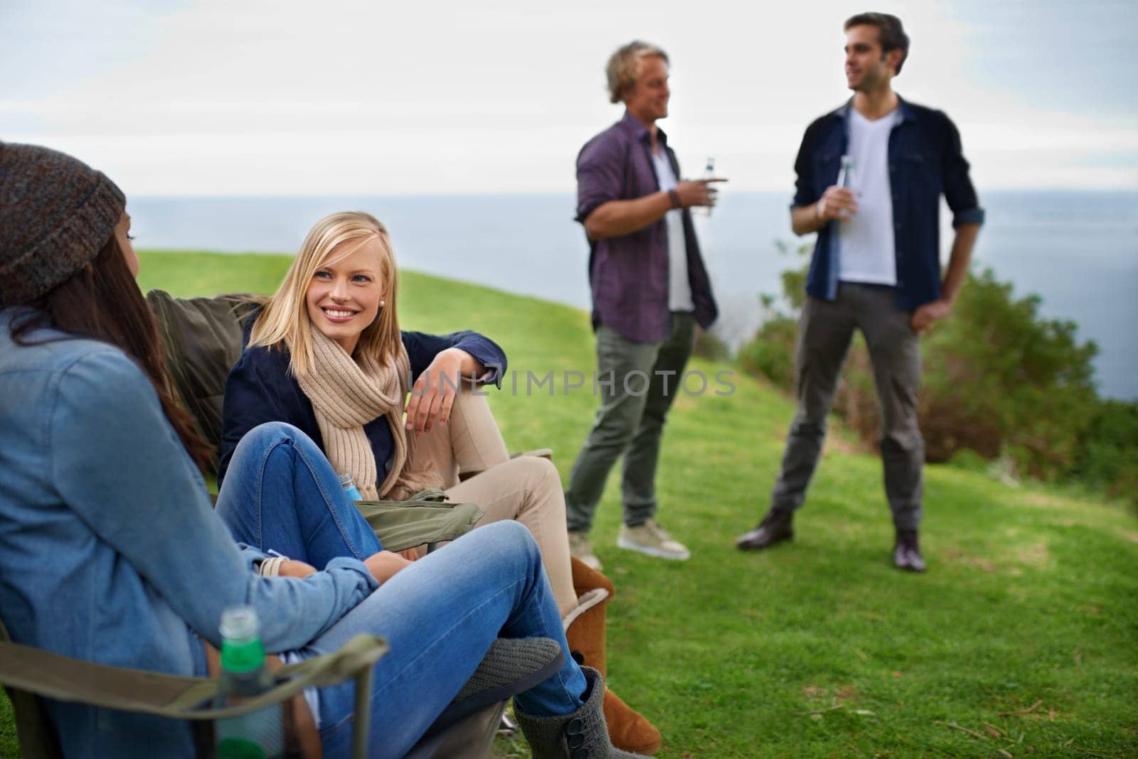 Friends, men and women with drinks for relax on grass in nature with happiness for holiday and vacation. People, smile and alcohol in countryside or lake for adventure, travel and bonding in forest by YuriArcurs