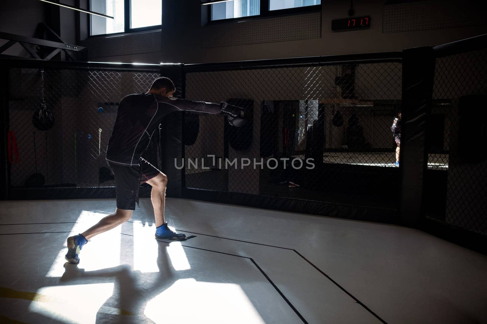 Dedicated male athlete engaging in shadowboxing session, simulating combat moves against imaginary opponent in boxing octagon cage in modern gym setting