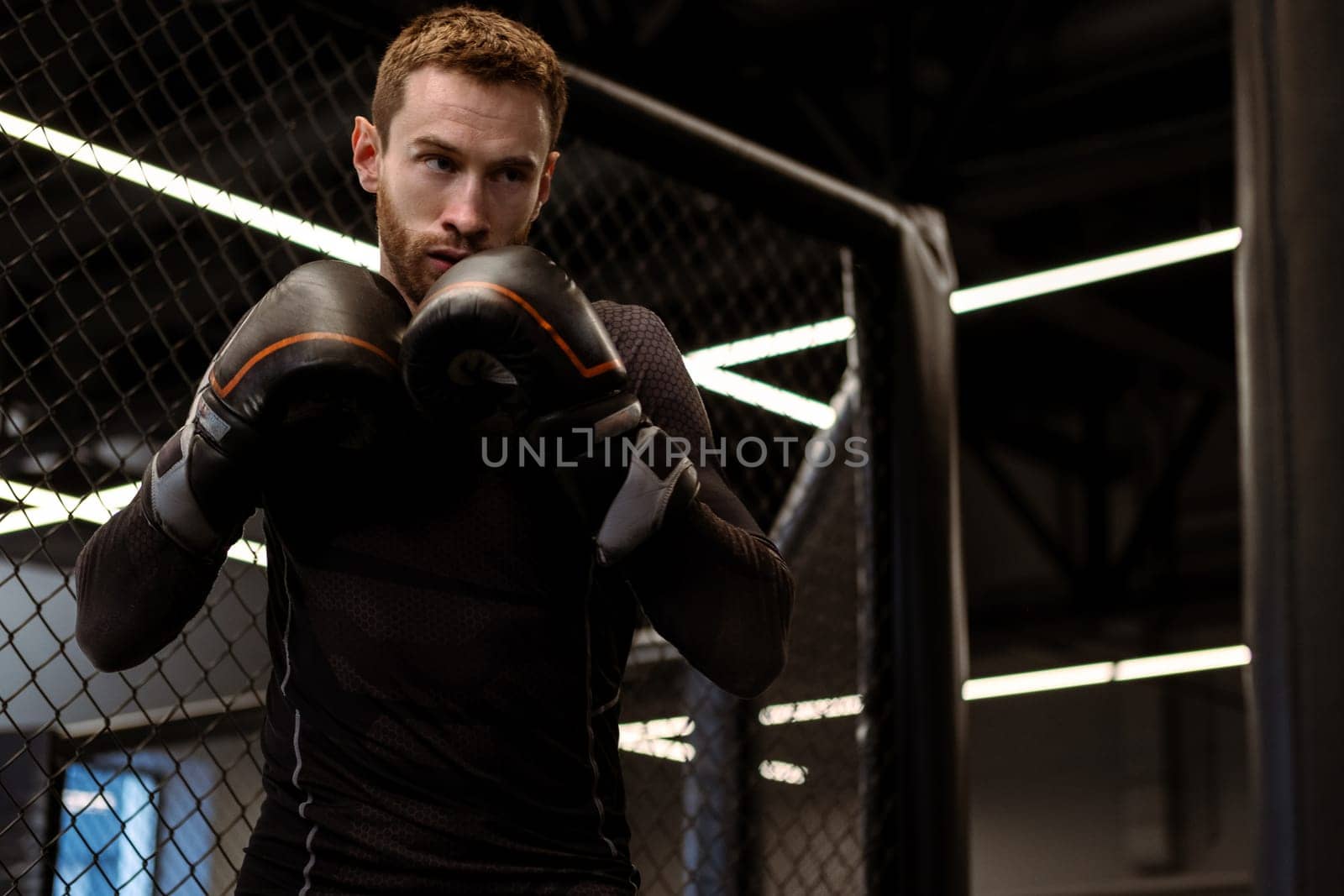 Concentrated young adult boxer wearing gloves standing in fighting stance, ready to throw powerful punch to heavy bag during training in modern boxing gym