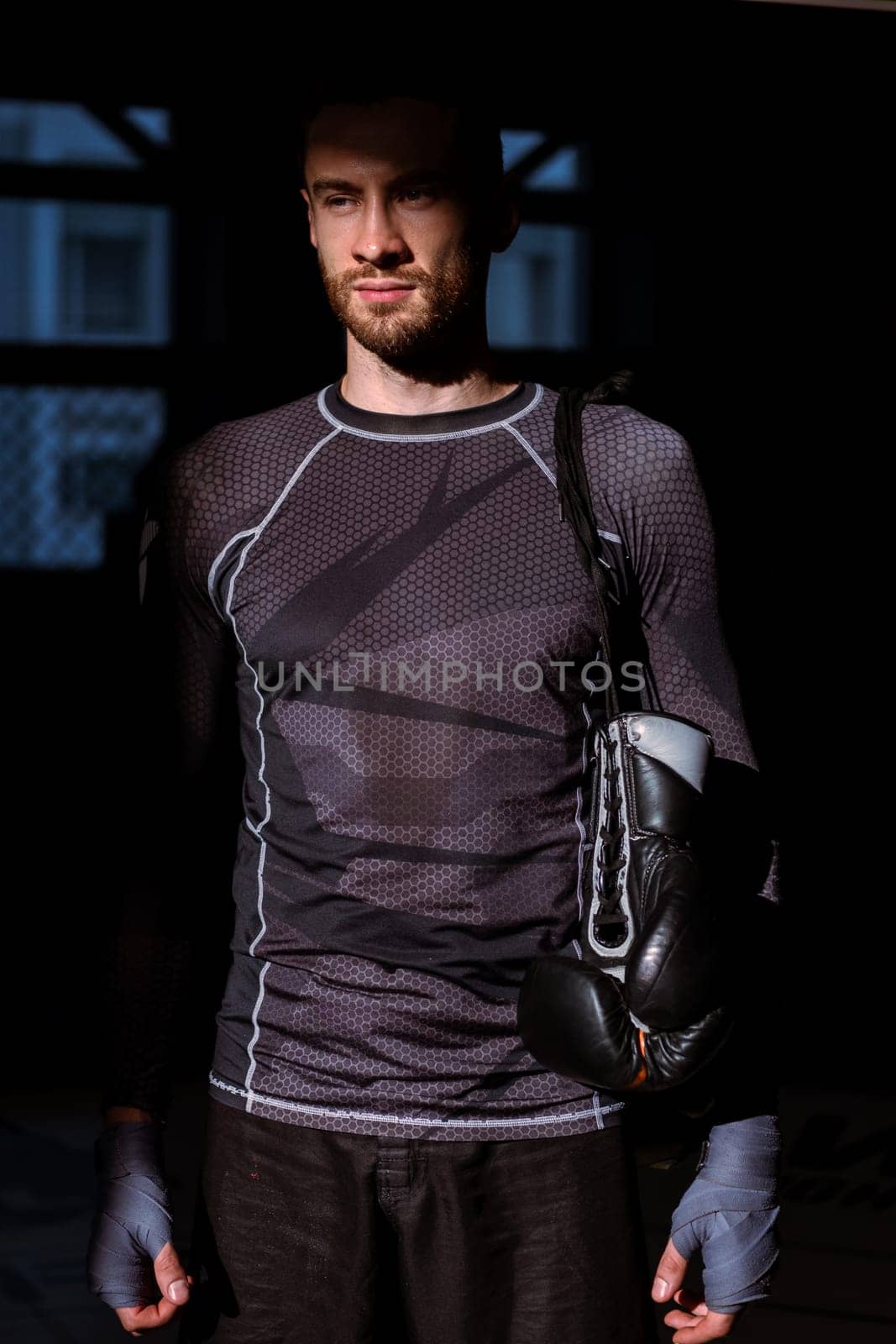 Confident relaxed bearded fighter with wrists wrapped in handwraps and gloves hanging over shoulder, standing in rays of sunlight in dark gym after successful training