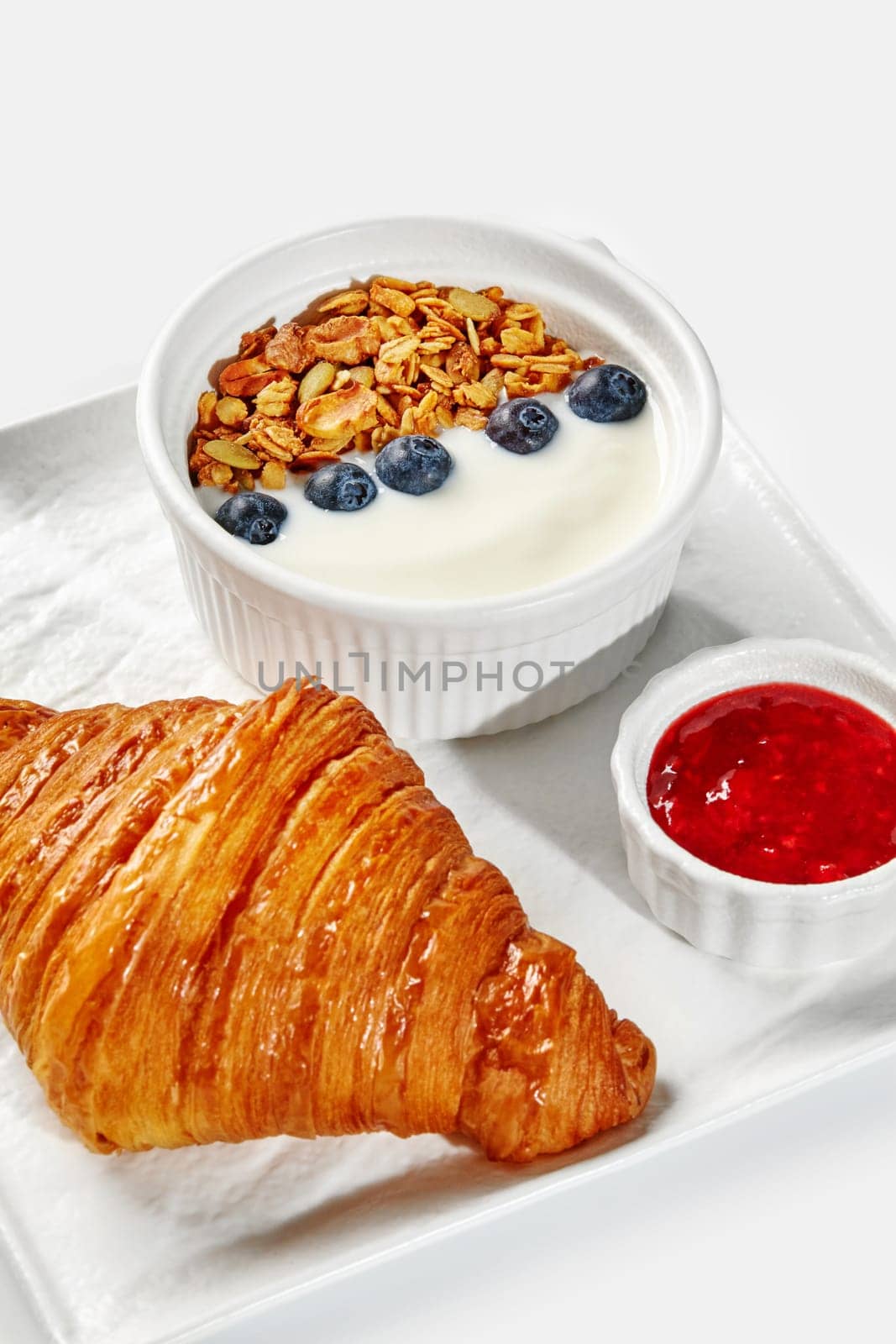 Delicious continental breakfast setup featuring buttery croissant, yogurt topped with granola and blueberries, and side of red berry jam served on white tray