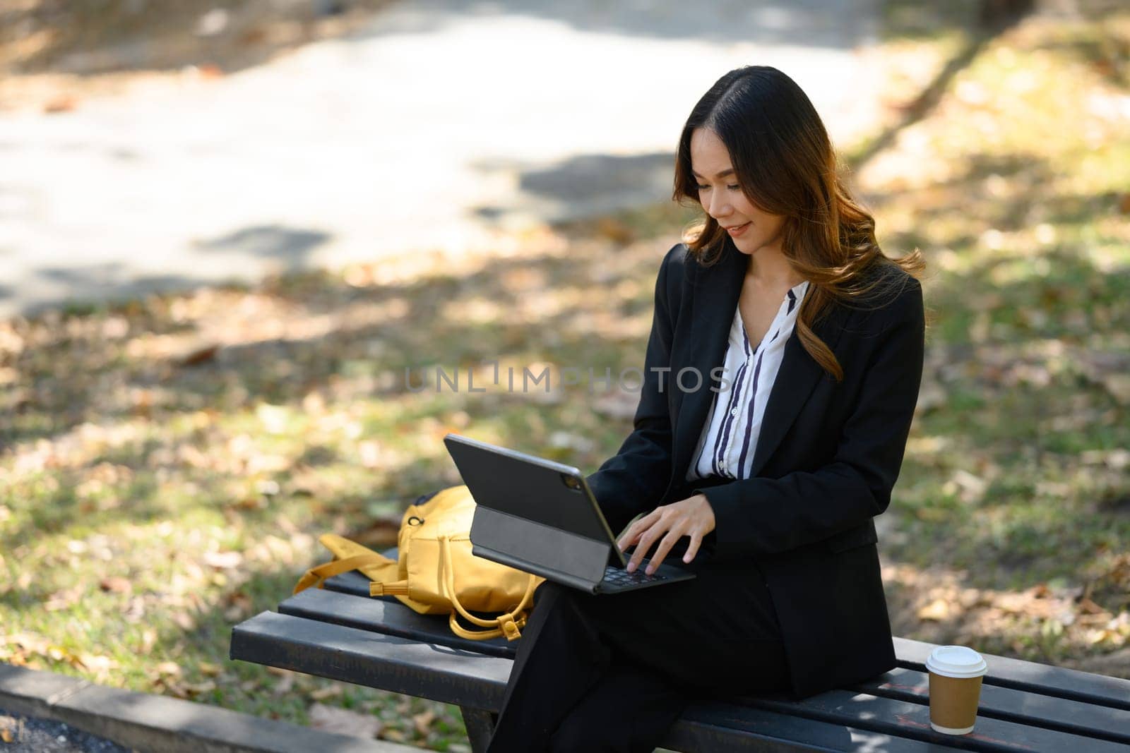 Charming businesswoman sitting on the bench in the city park and using digital tablet.