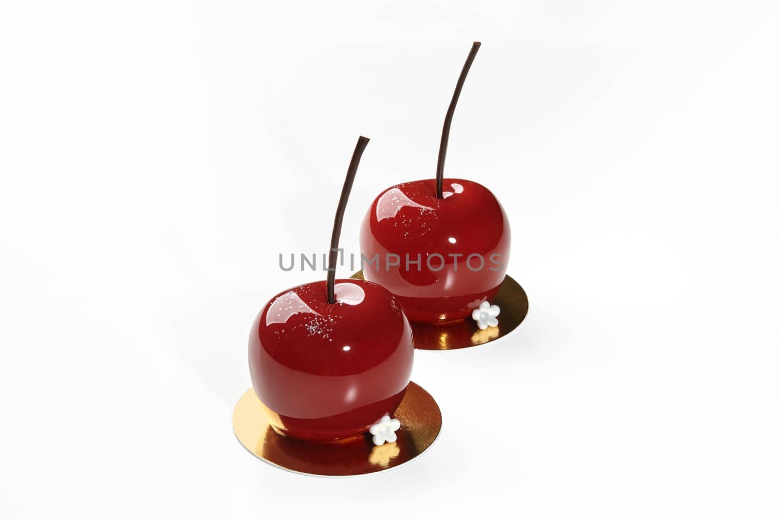 Two exquisite cherry-shaped desserts with mirror-like red glaze and delicate chocolate stems, presented on golden serving cardboards on white