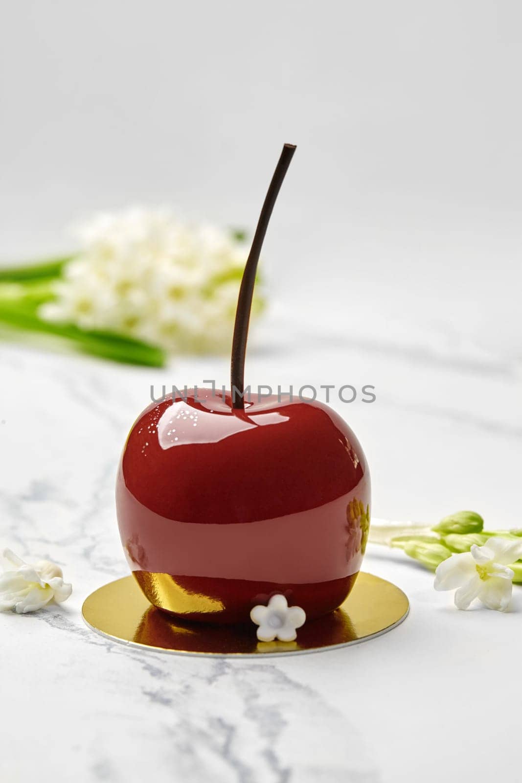 Artisan cherry-shaped dessert, inspired by nature simplicity, with vibrant red glaze and chocolate stem served on gold cardboard pastry base, on marble top with sprig of white flowers