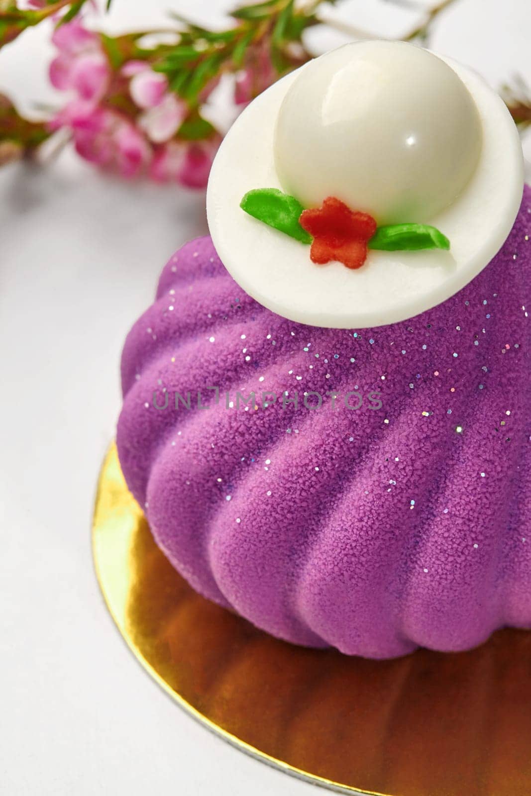 Close-up of velvety texture of luxurious purple mousse pastry topped with white chocolate hat on gold plate, on blurred background with vibrant pink flowers