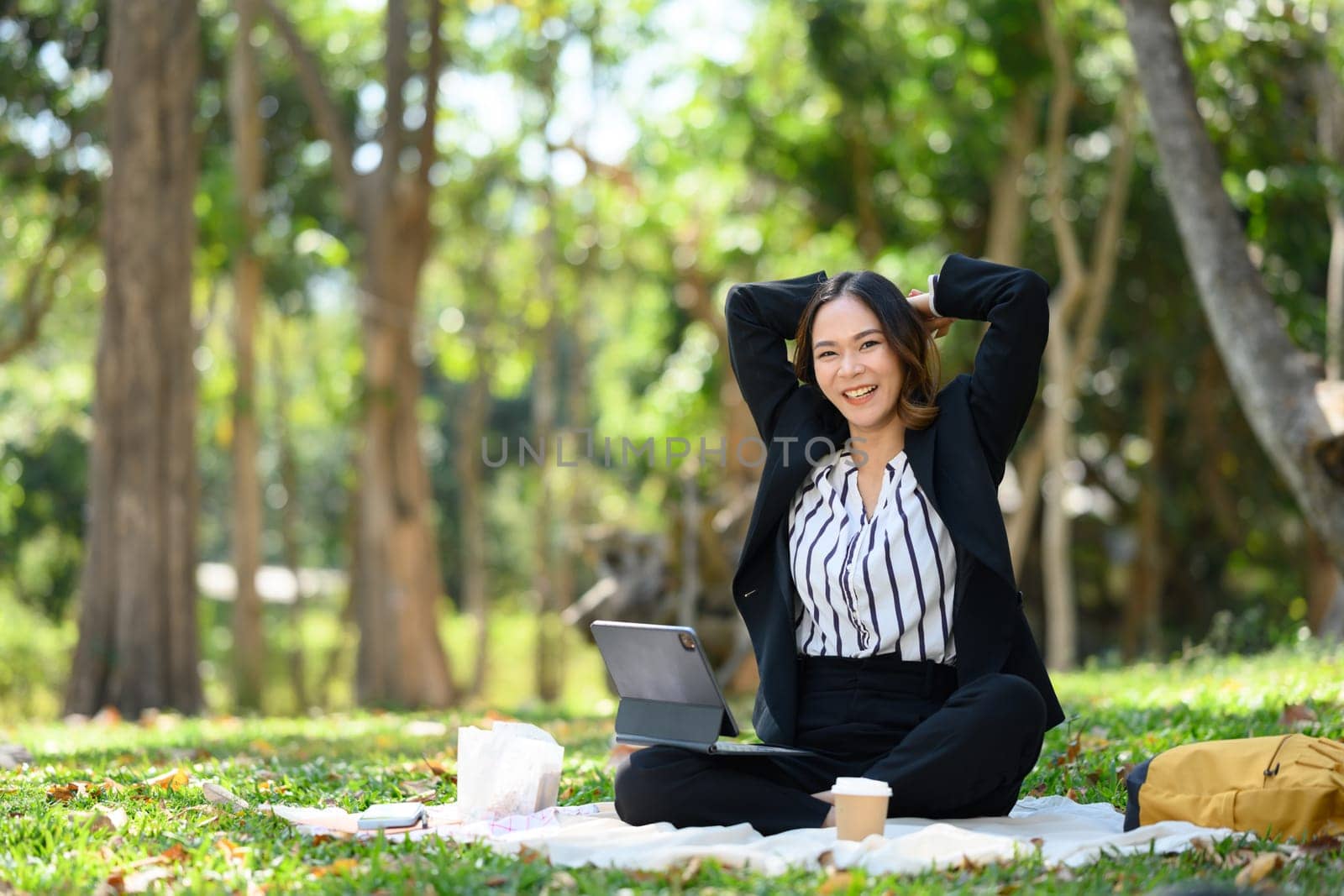 Happy millennial businesswoman sitting with digital tablet on green grass and stretching arms.