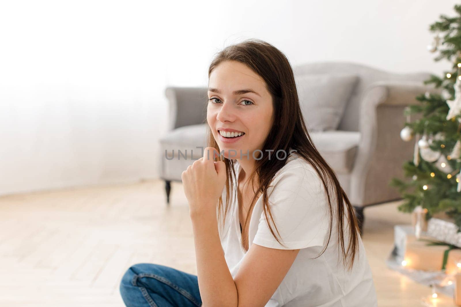 Beautiful young brunette in casual outfit sitting on floor at home and looking at camera on background of fir tree