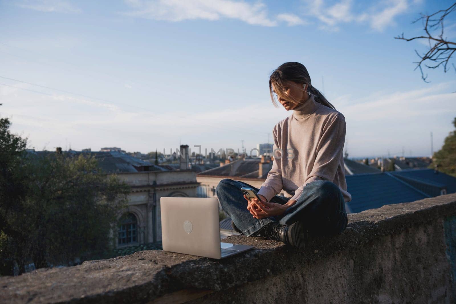 Woman freelancer uses laptop on cement wall outdoors against the sky and the roof of the city. The woman to be focused on her work or enjoying some leisure time while using her laptop. by Matiunina