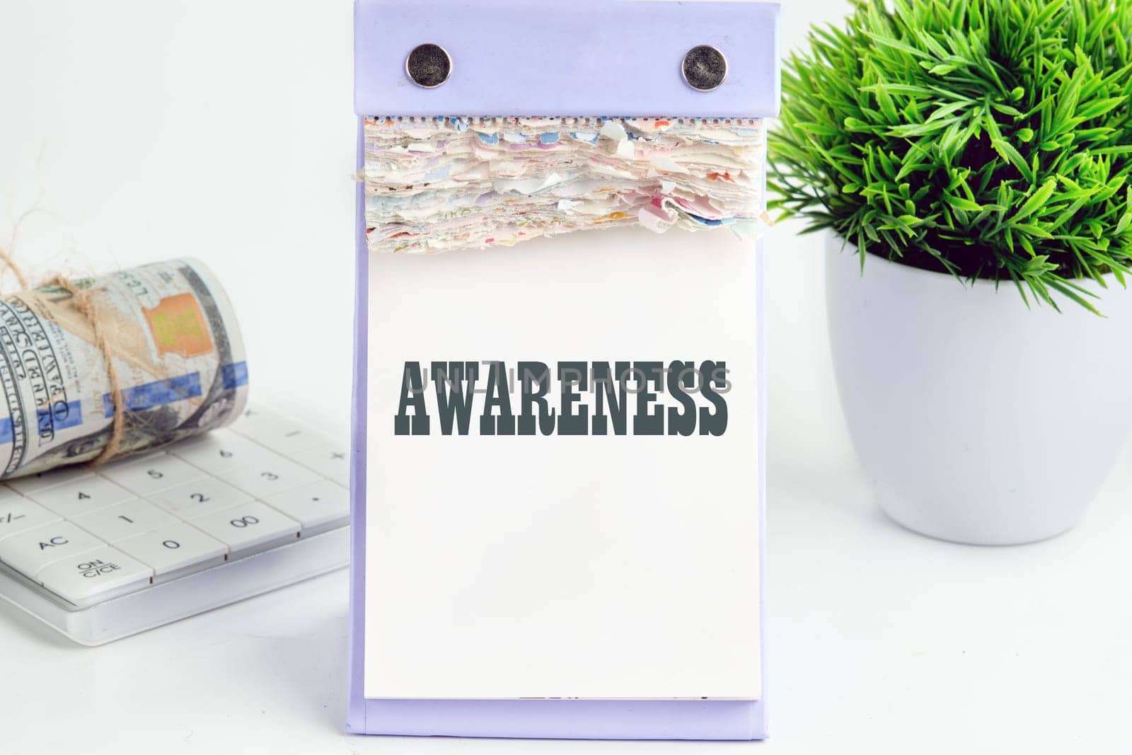 AWARENESS word writing on a desktop tear-off calendar on a white background, next to a calculator with a roll of banknotes with a flower out of focus in the background
