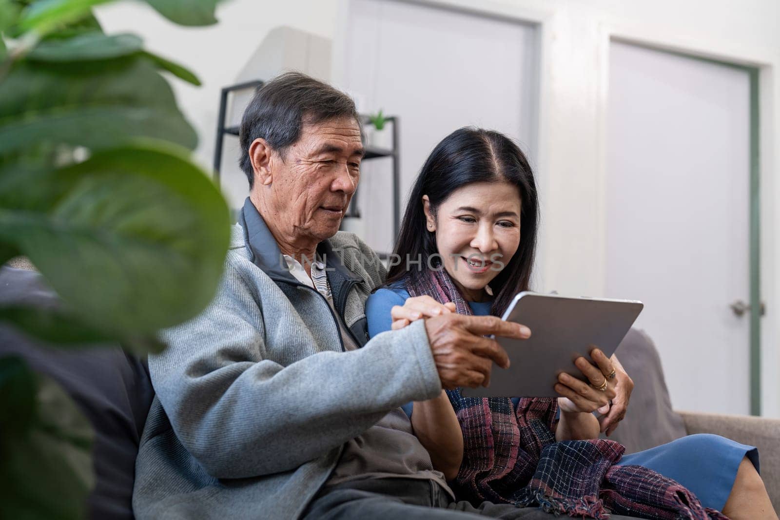 Happy elderly asian couple using tablet sit on sofa doing ecommerce shopping online on website and buying insurance browsing at home.