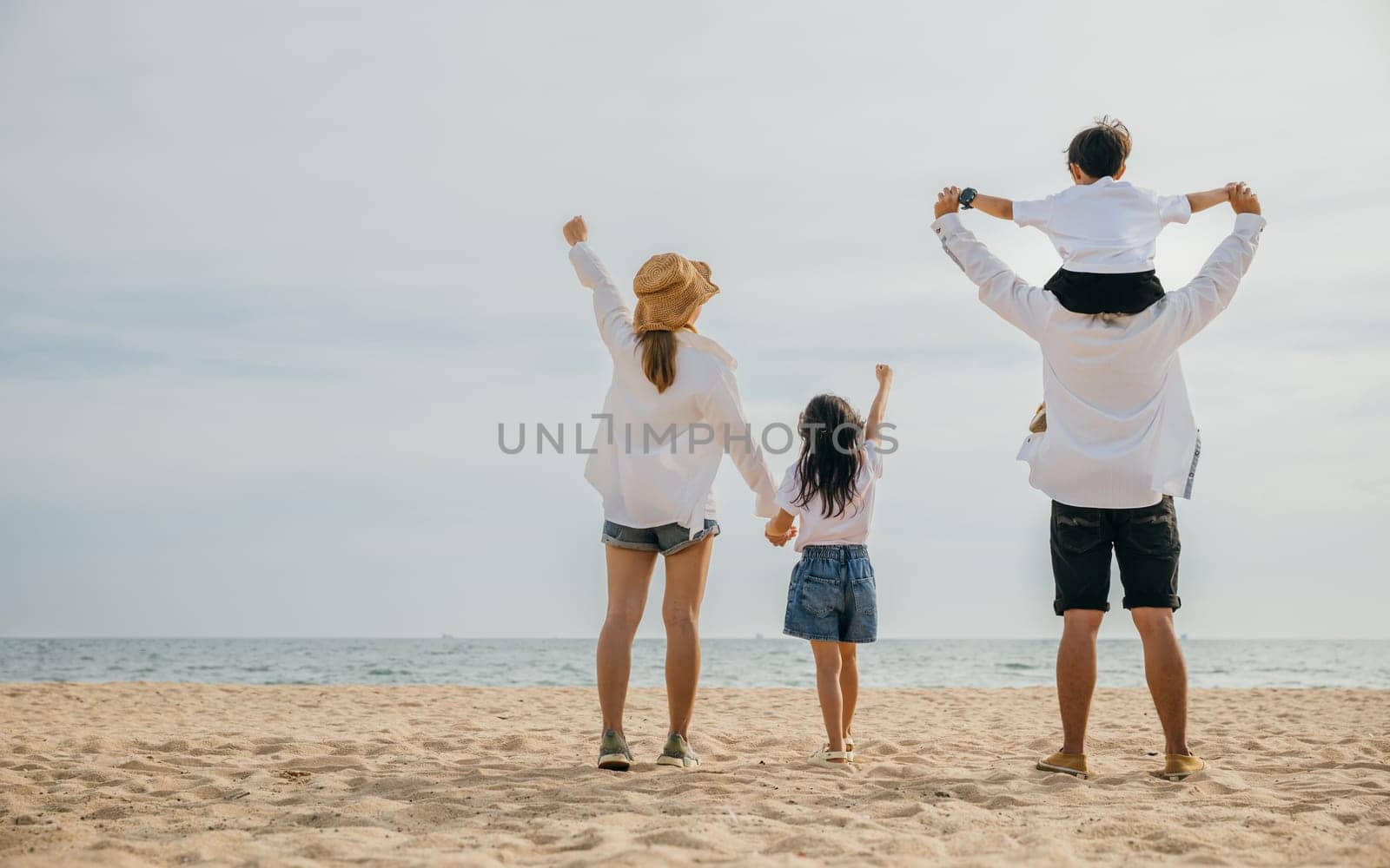 A joyful family of four stands with raised arms at the sea beach during sunset. Father carries his son on shoulders capturing the carefree moments of a delightful summer vacation. by Sorapop
