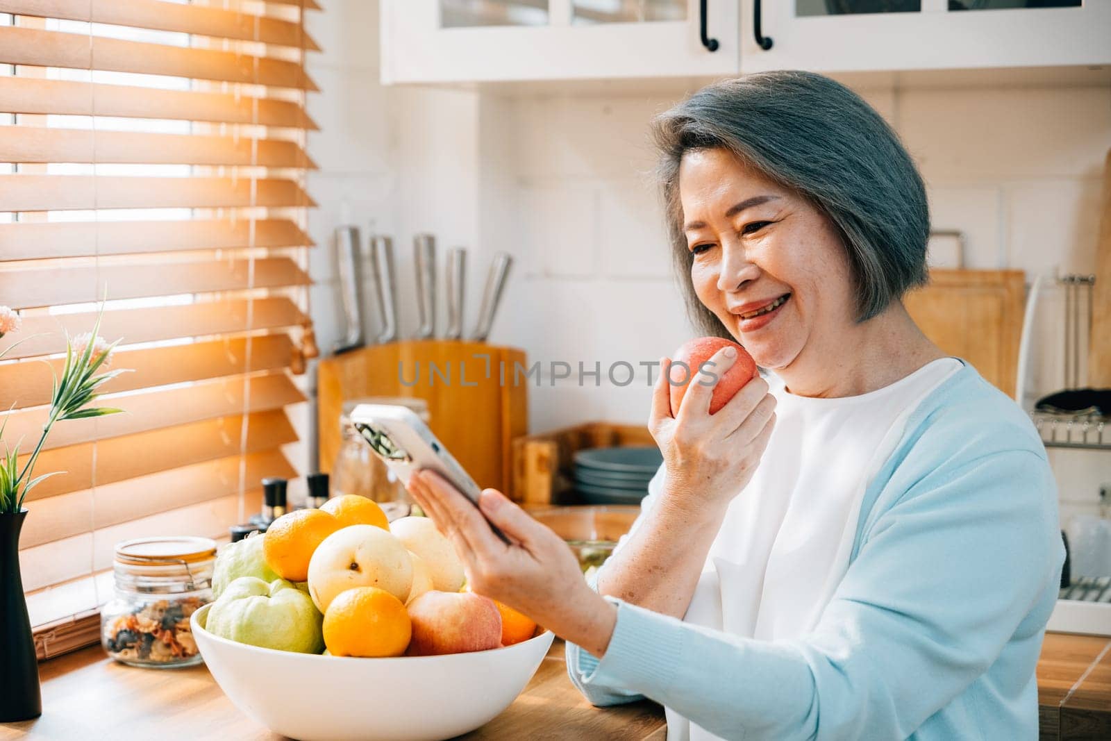 In the kitchen, an old woman, a grandmother, savors breakfast. She's smiling, using her smartphone, and munching on a red apple. A portrayal of technology, happiness, and care at home.