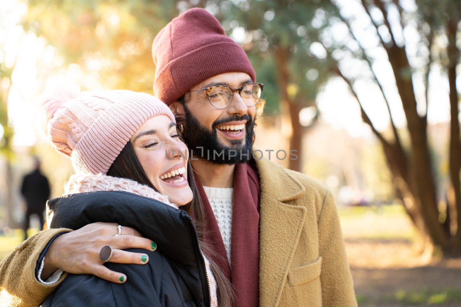 A man with a beard and wearing outerwear stands next to a happy woman in a park. He gestures with his finger, both wearing headgear and smiling