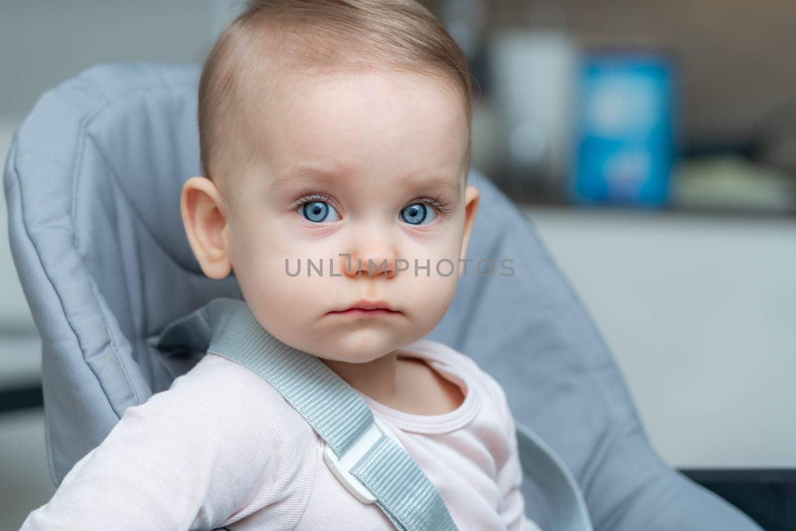 A baby with adorable cheeks and a cute hairstyle is sitting in a seat, looking directly at the camera with their bright iris and long eyelashes