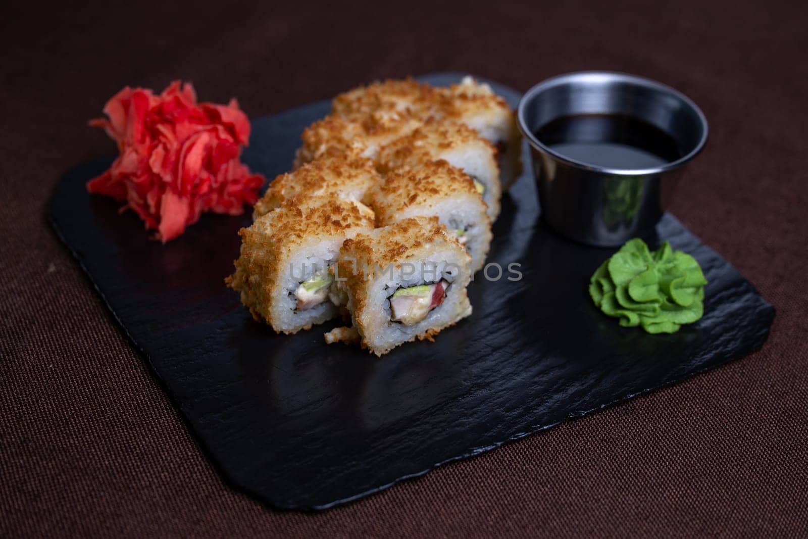 Deep-fried sushi roll on black stone plate with soy sauce, wasabi. Made with rice, seaweed, vegetables, shrimp tempura.