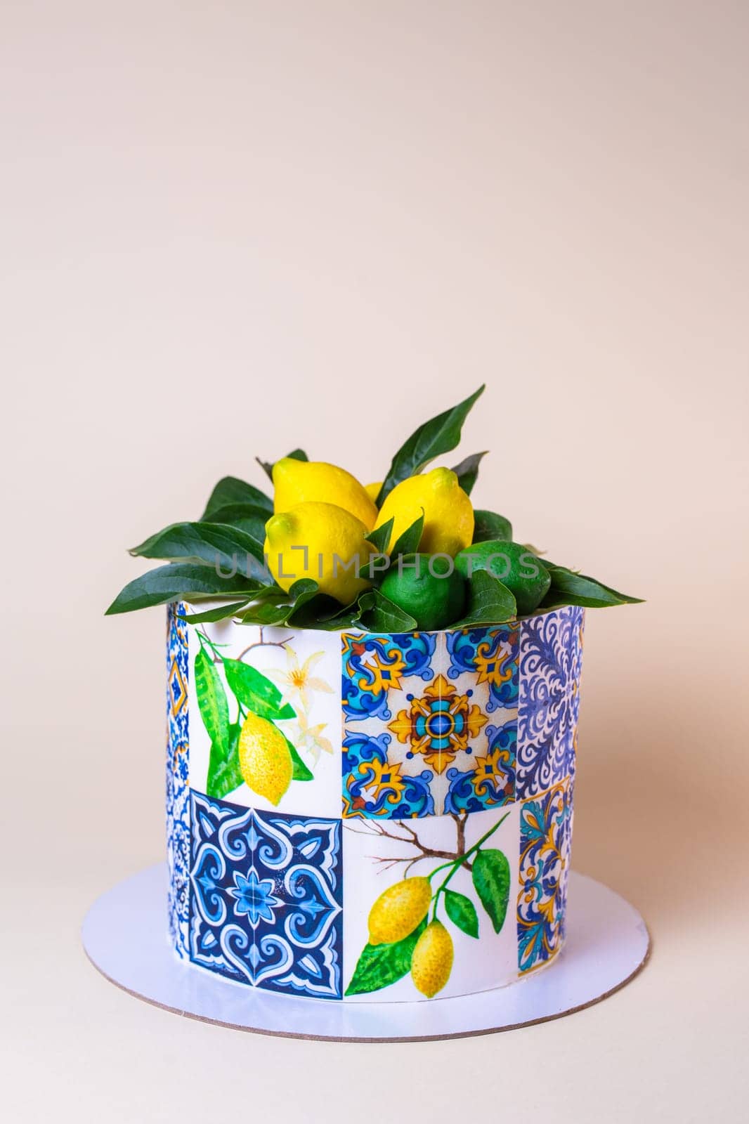 A round cake is decorated with lemons, green leaves, and ceramic tiles pattern. The cake is isolated on a beige background.