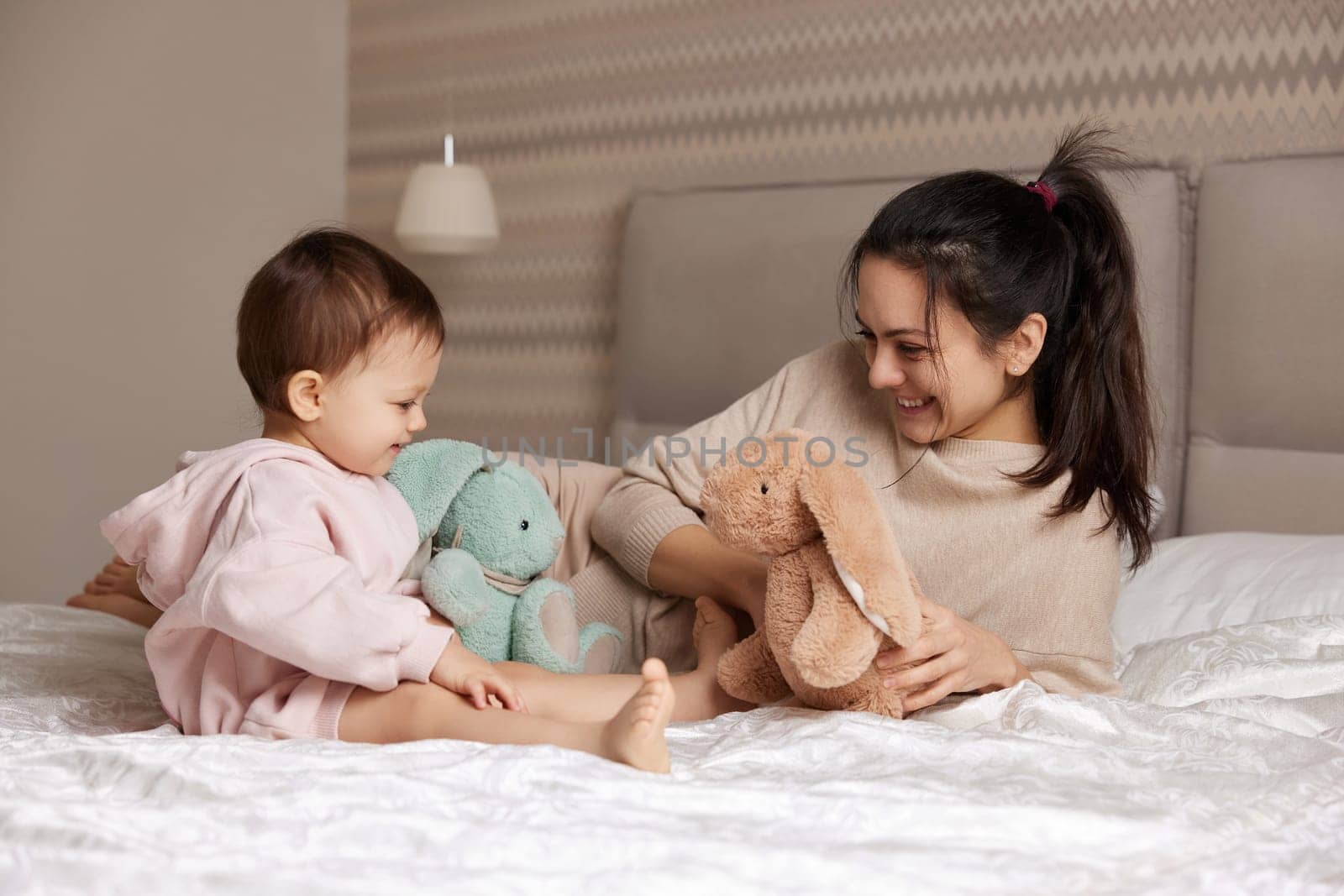happy mother and her little child daughter playing with bunny toys in bedroom, family having fun