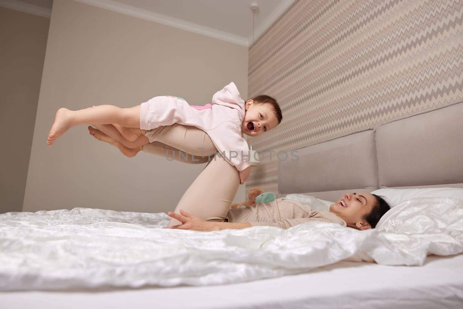 Happy Caucasian mother raising laughing cute baby and playing together in bedroom