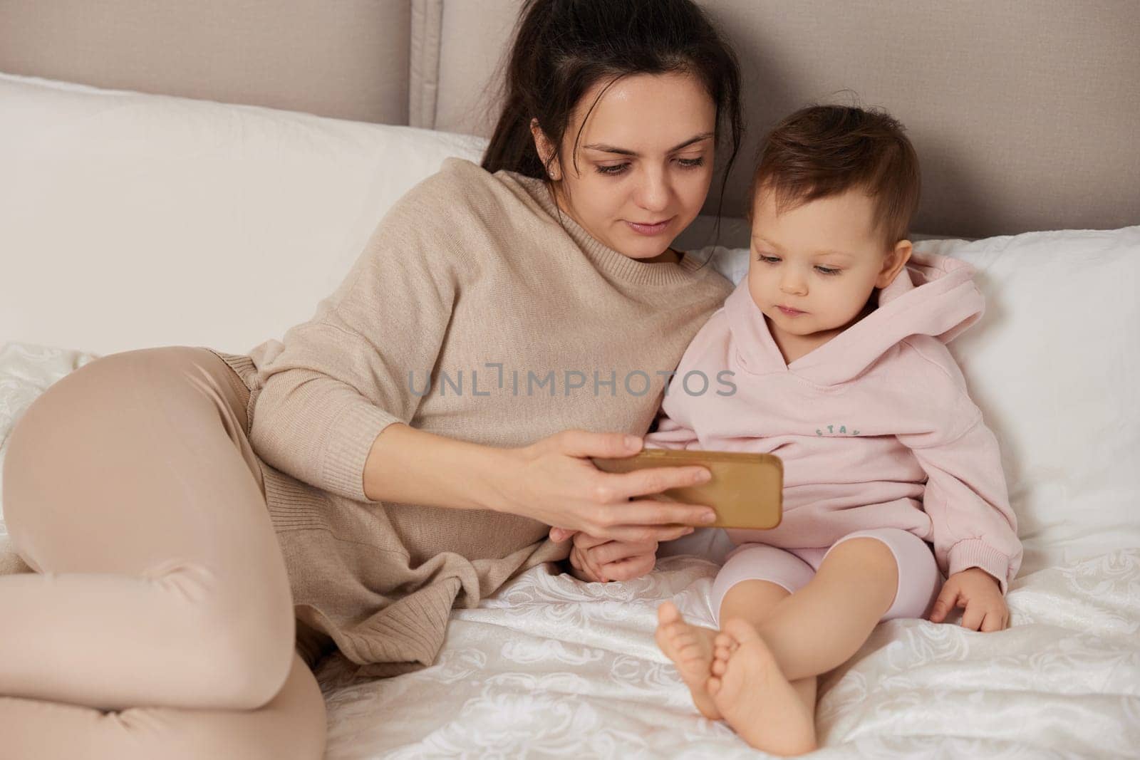 Caucasian mother and child playing with smartphone while sitting on bed in bedroom at home