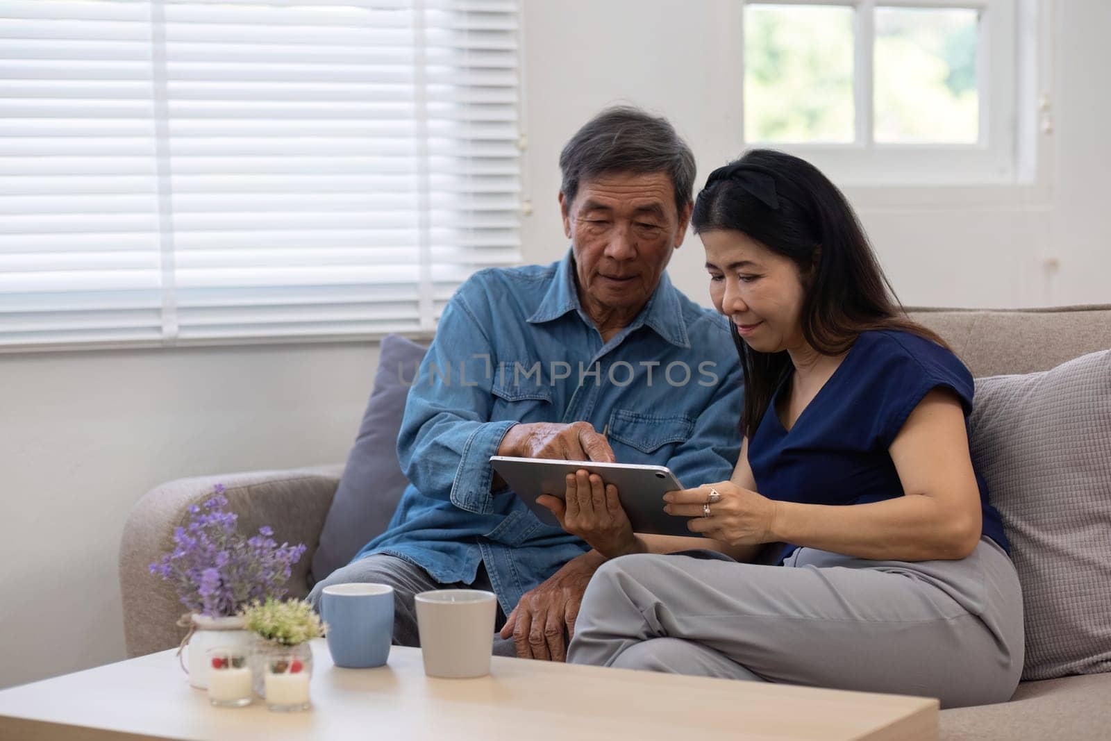 Cheerful Asian couple using network video call tablet sitting together on sofa at home Asian elderly husband sitting with elderly wife shopping online or video calling on tablet, relaxing. by wichayada